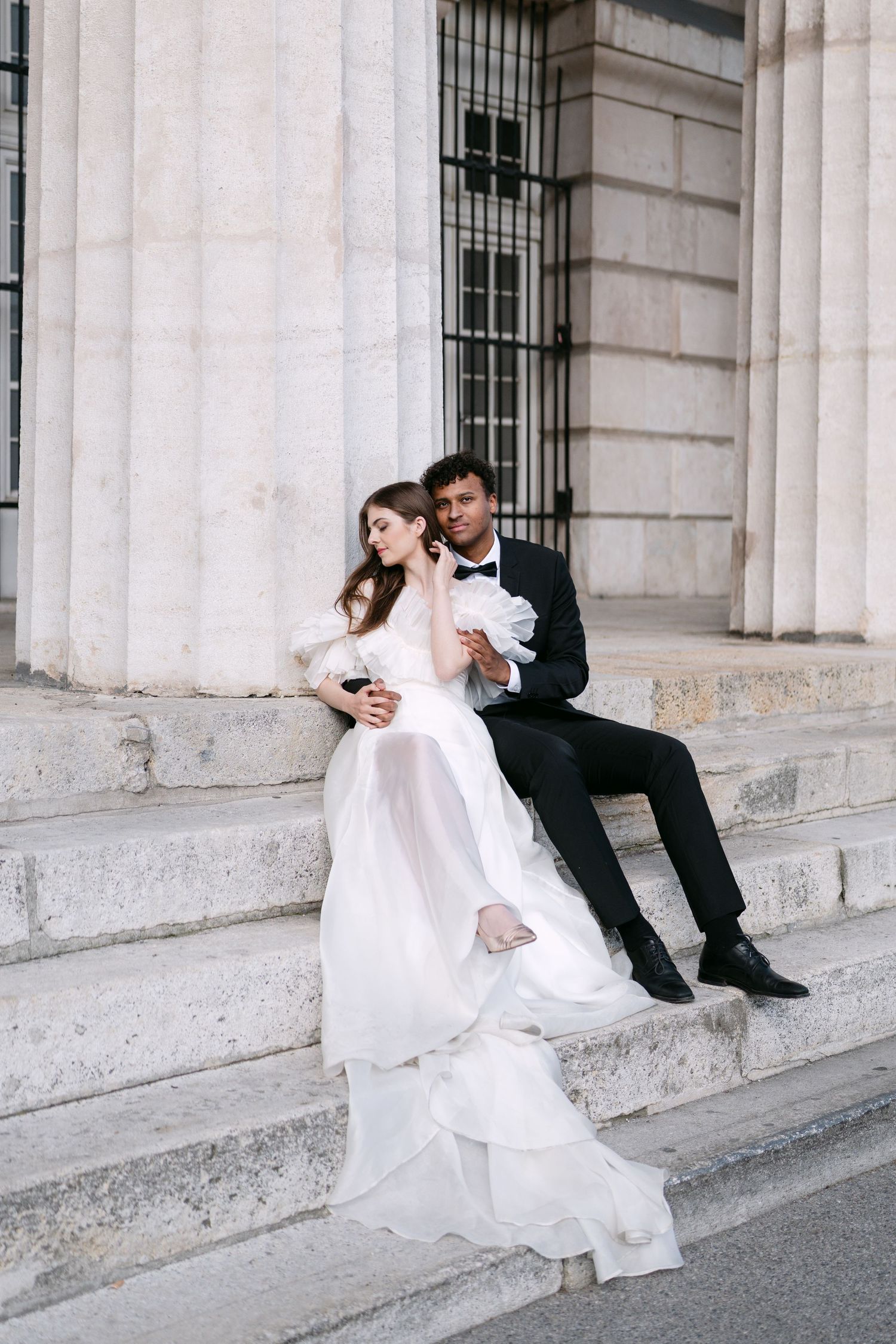 Wedding couple relaxes on classical stone steps in formal attire.