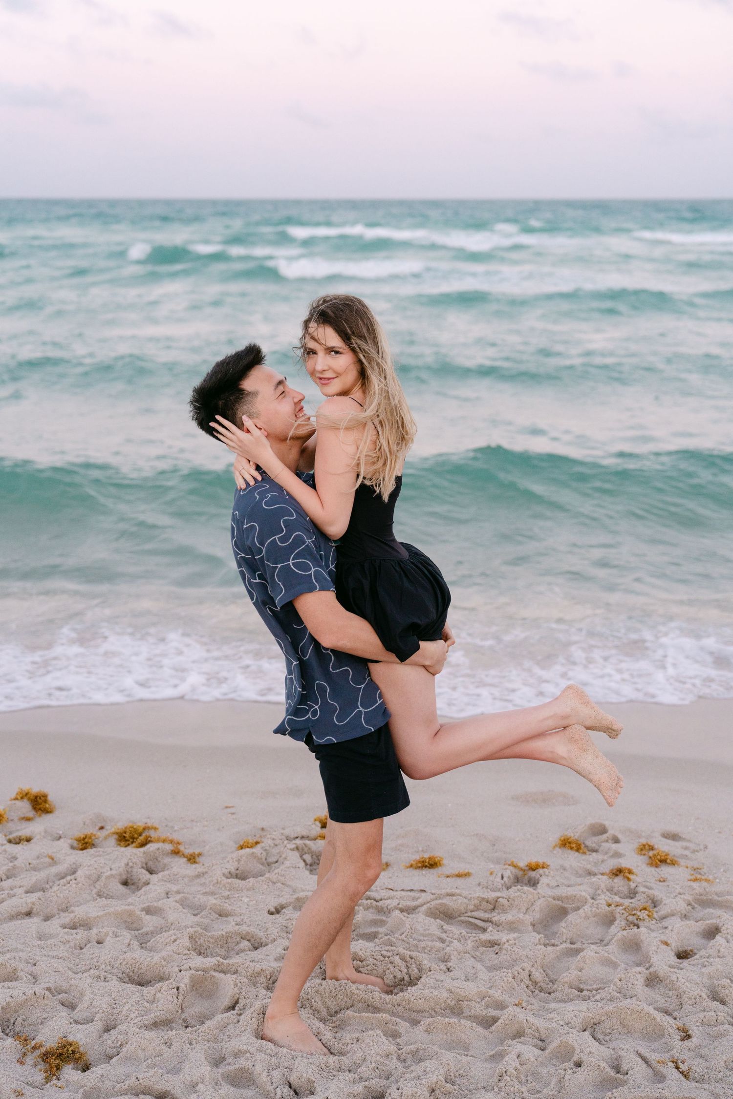Romantic couple embraces and plays on a sandy beach with turquoise ocean waves in the background.