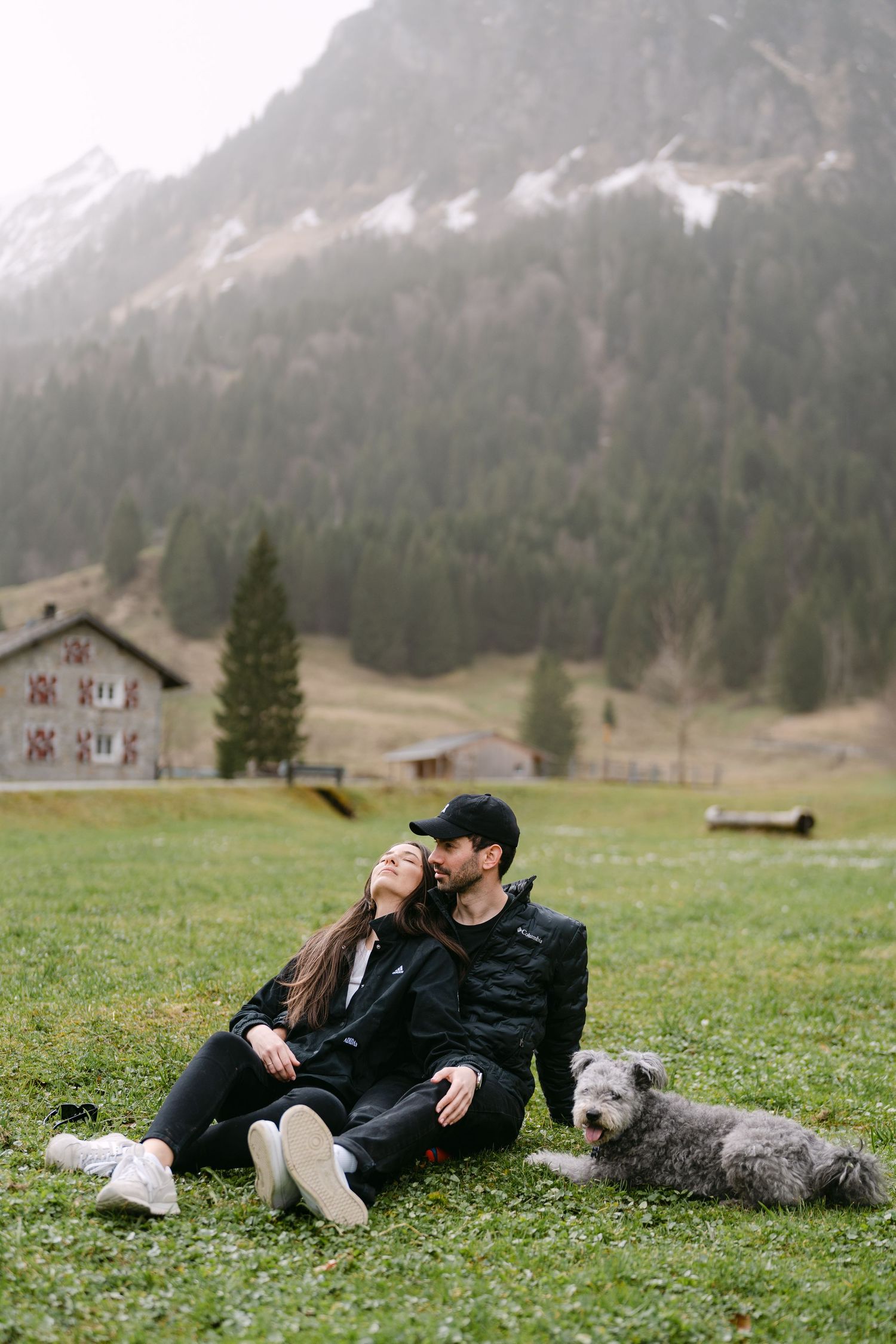 Couple relaxing on grass with their dog against mountain scenery.