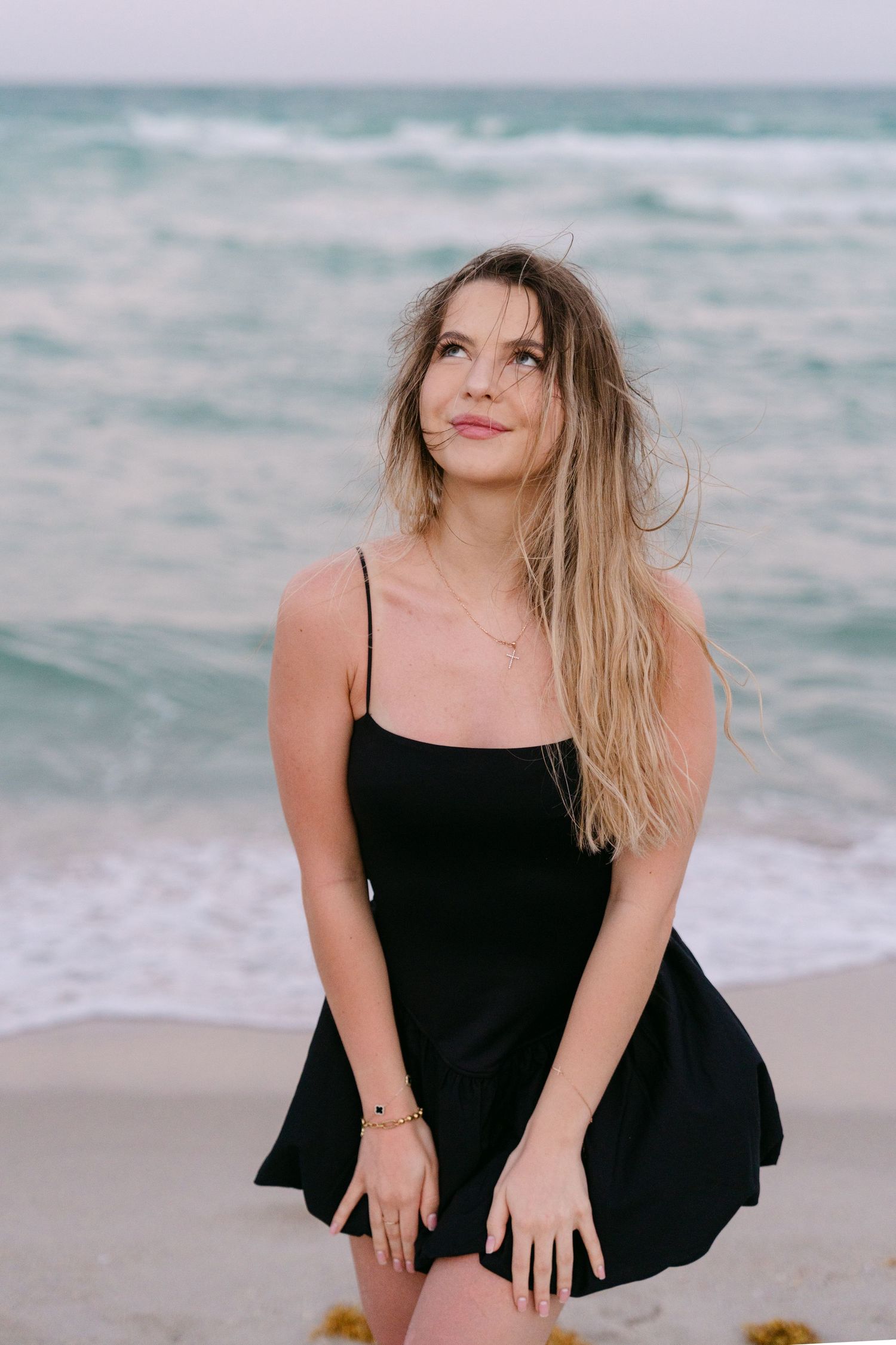 Woman in black dress poses playfully on sandy beach with ocean waves in background.