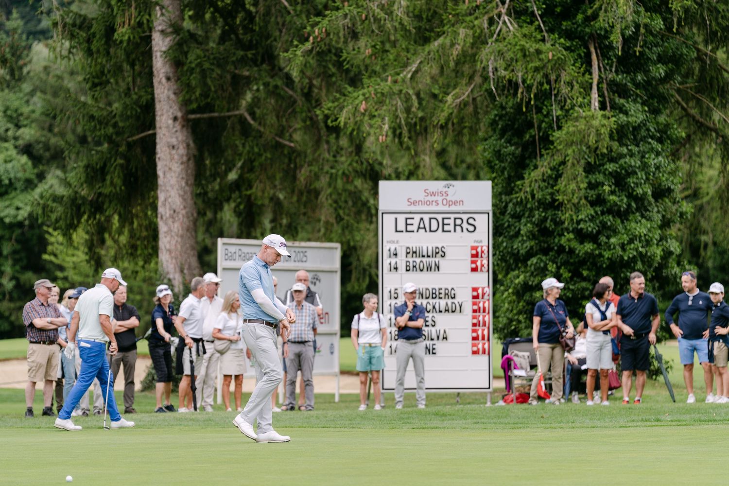 Wide view of golf tournament spectators and players on a tree-lined course.