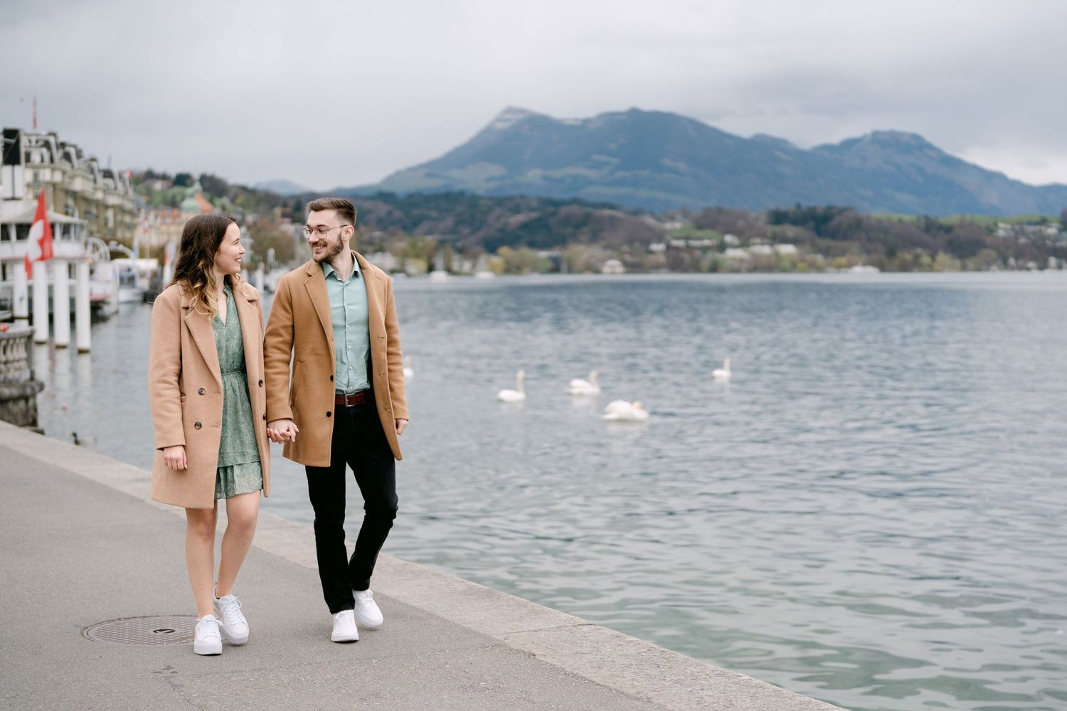 A couple strolls along a scenic waterfront with mountains and sailboats in the distance.
