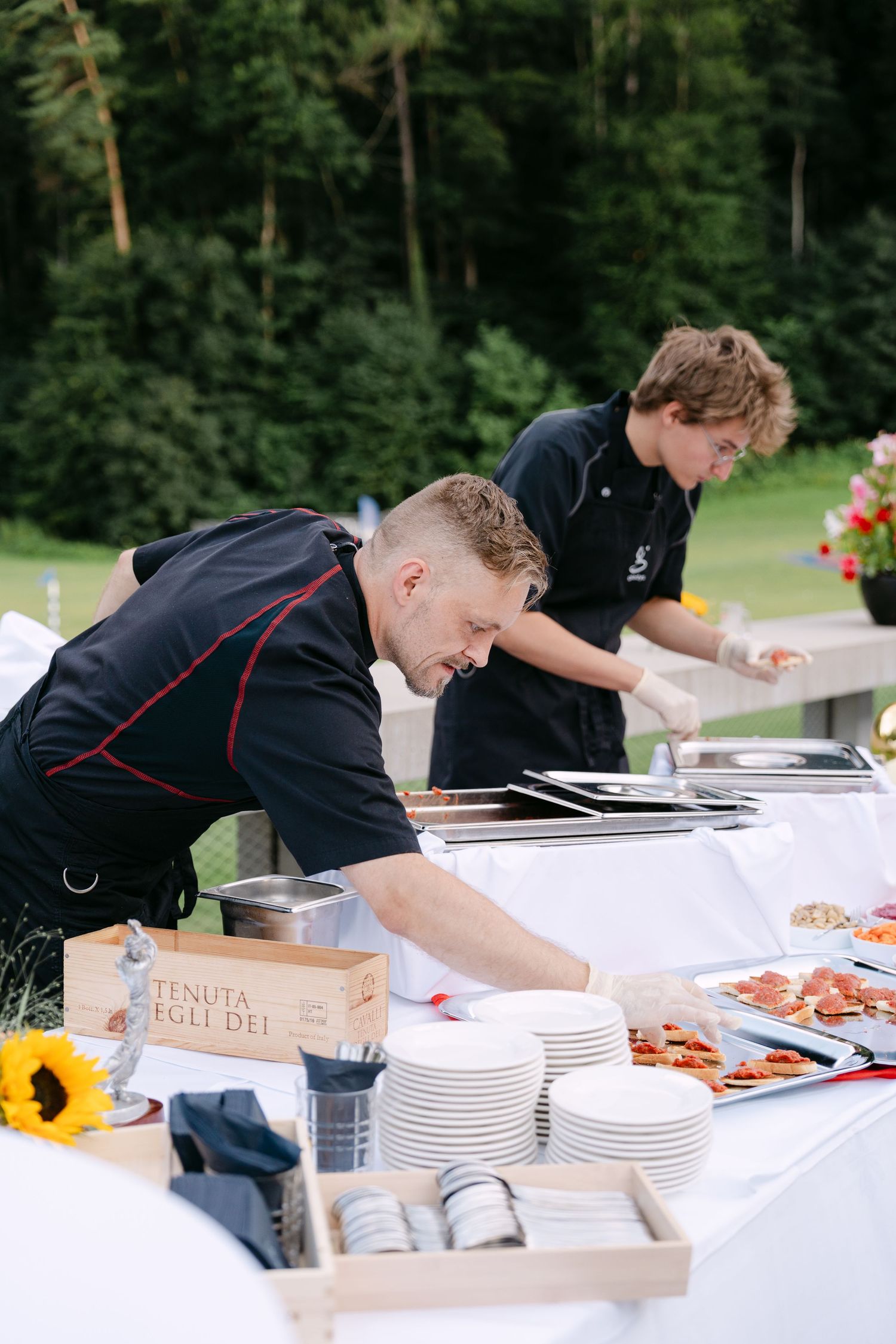 Catering team in black uniforms setting up plates and food stations at an outdoor event.