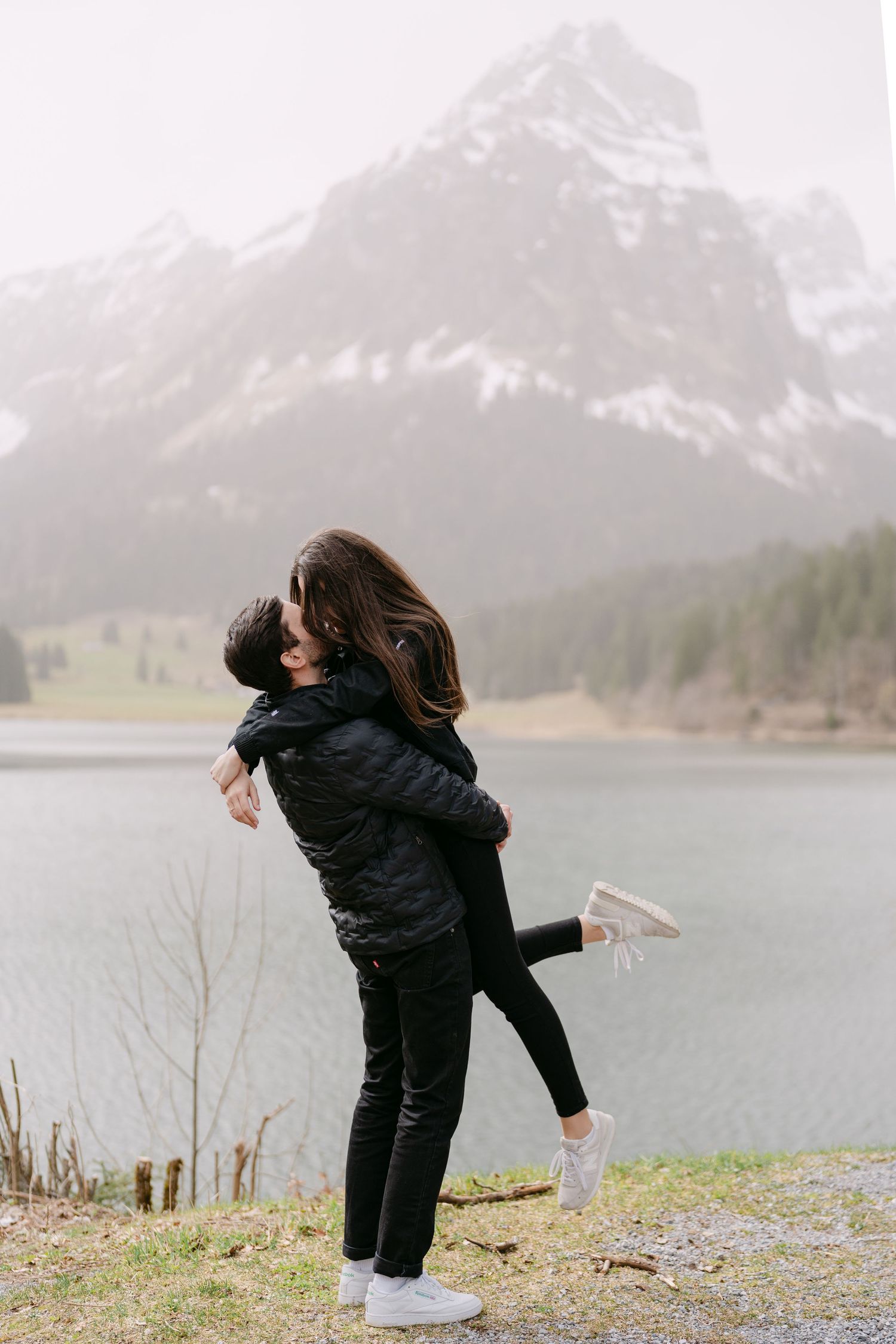 Playful couple in black outfits dancing by a mountain lake.