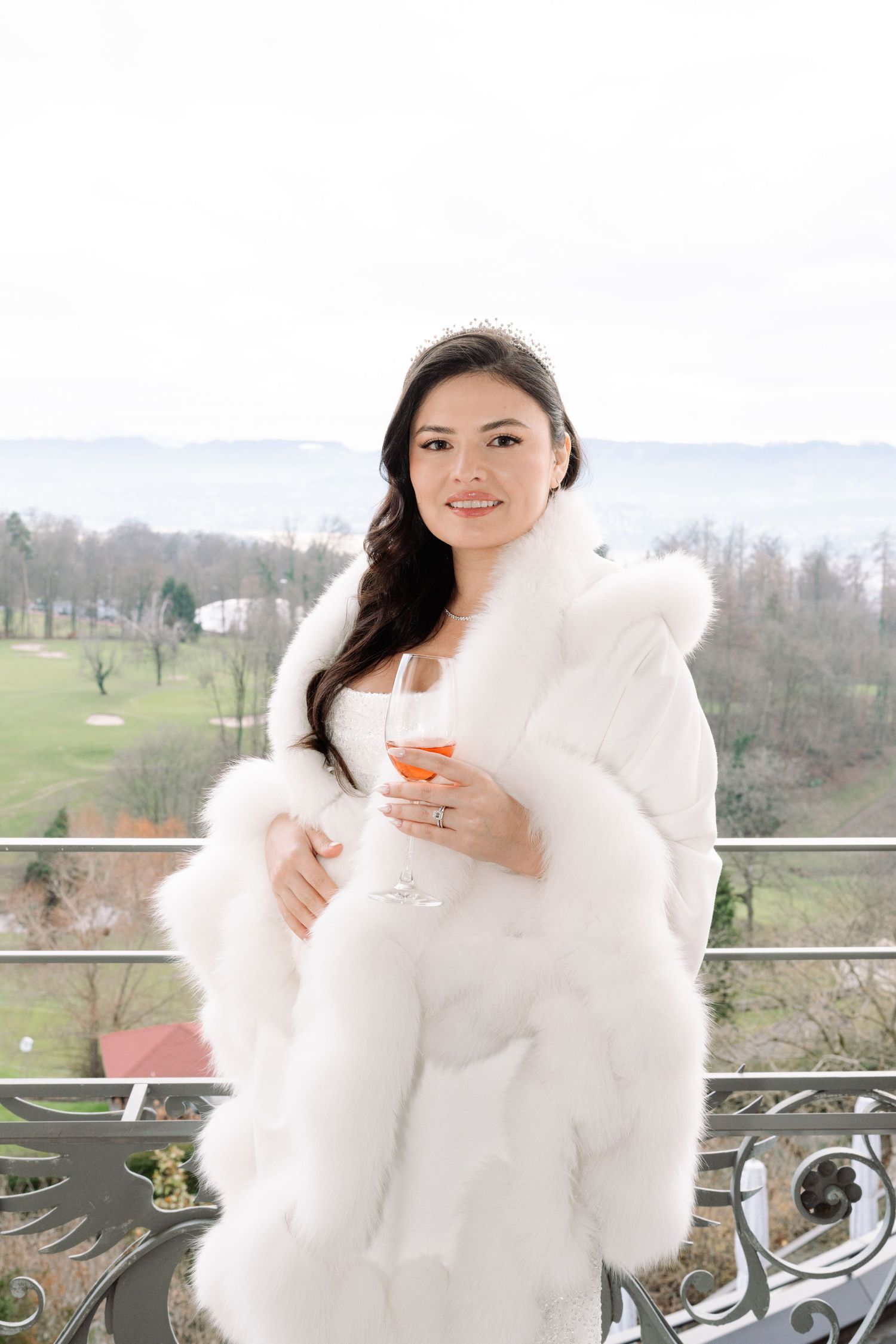 Person in white fur coat holds champagne on winter balcony with scenic view.