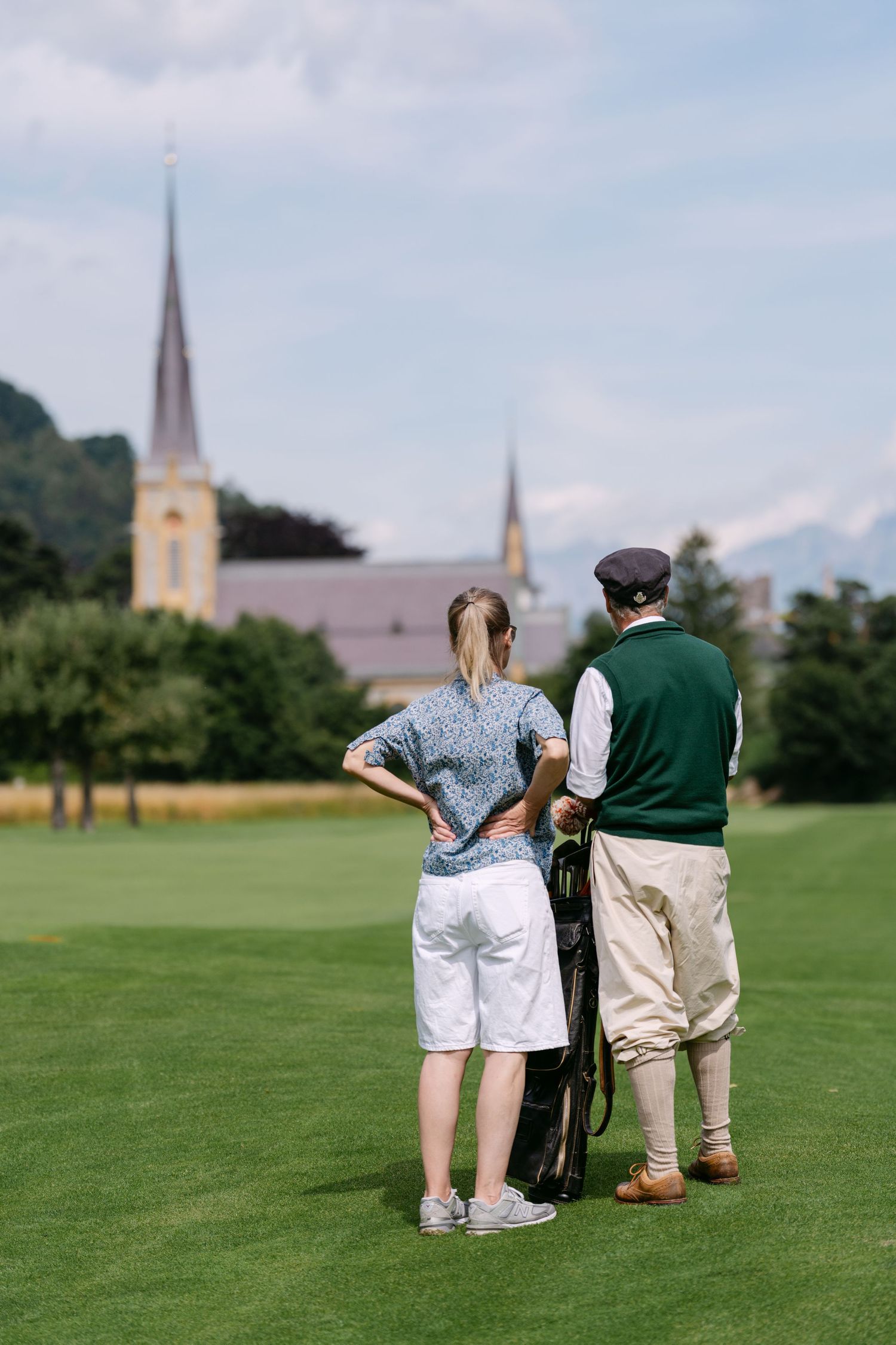 Players observing a croquet game on a well-maintained lawn with church buildings in the distance.