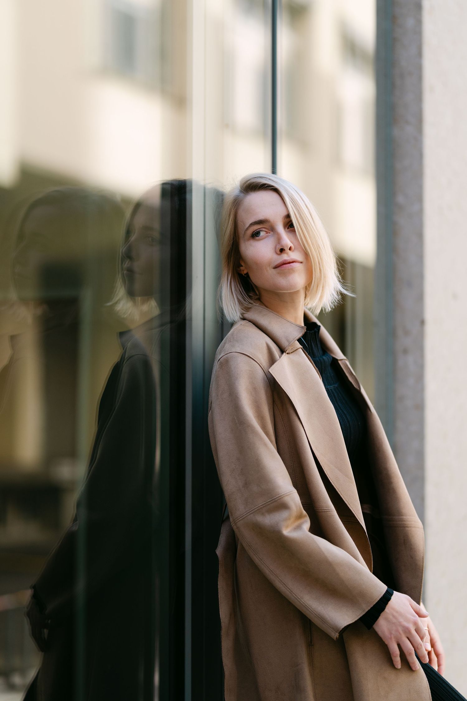 Casual portrait series showing someone in a tan coat posing near glass windows.