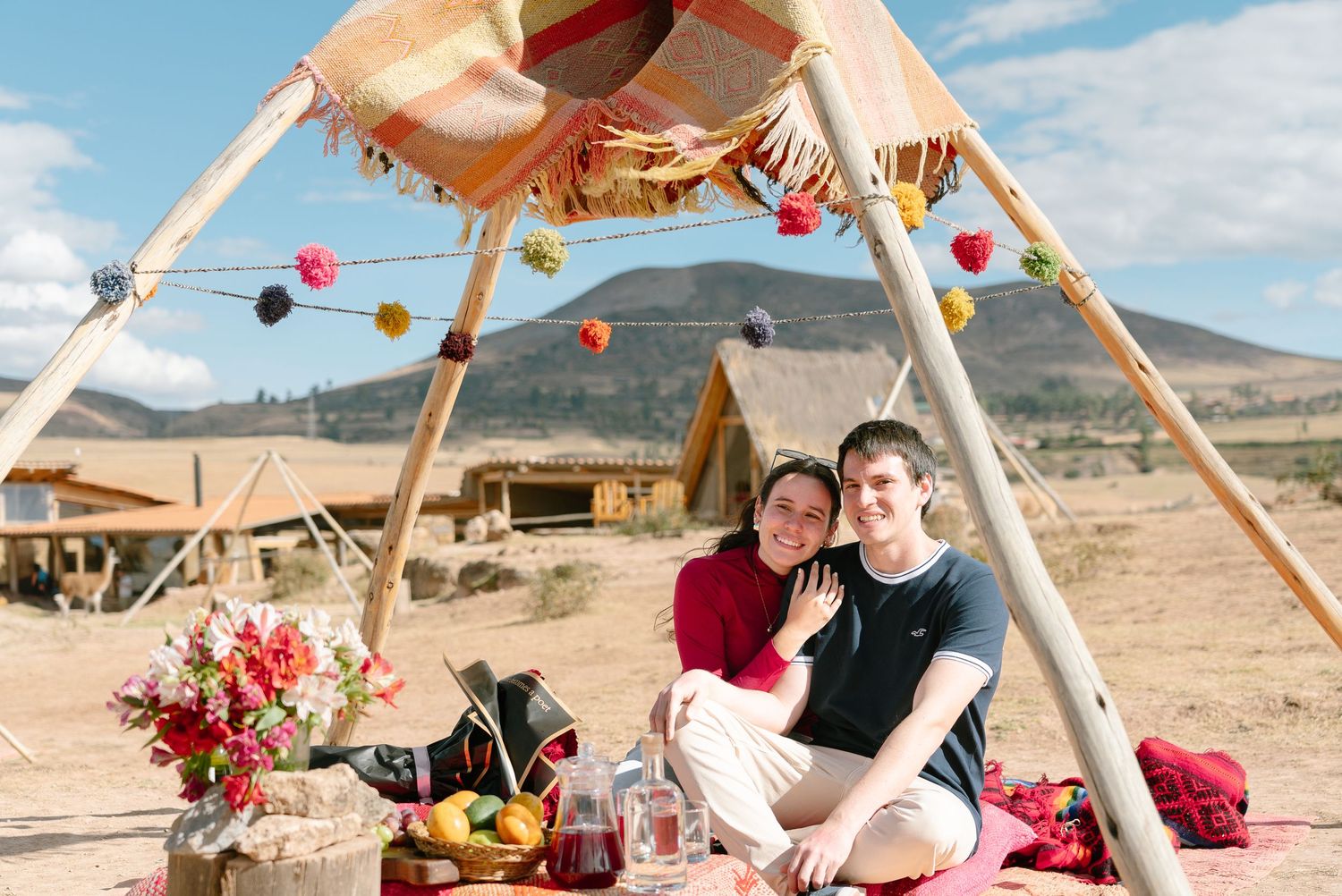 Desert picnic setting with wooden teepee structure decorated with colorful pom poms and string lights.