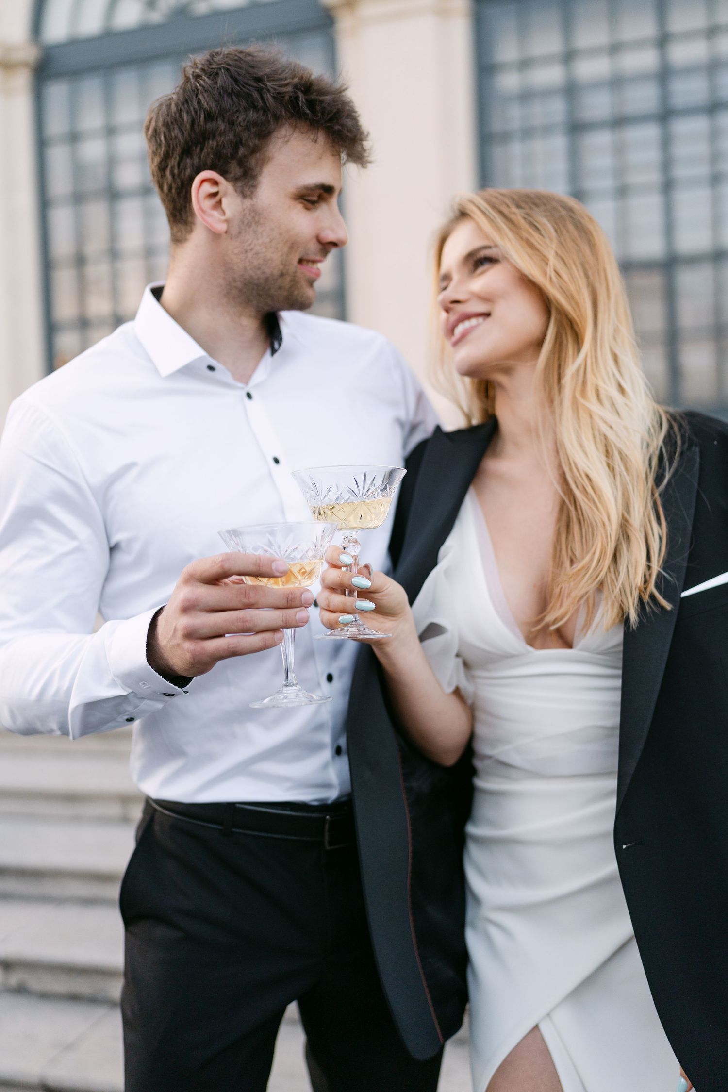 Stylish couple sharing champagne and laughter outside an elegant building.