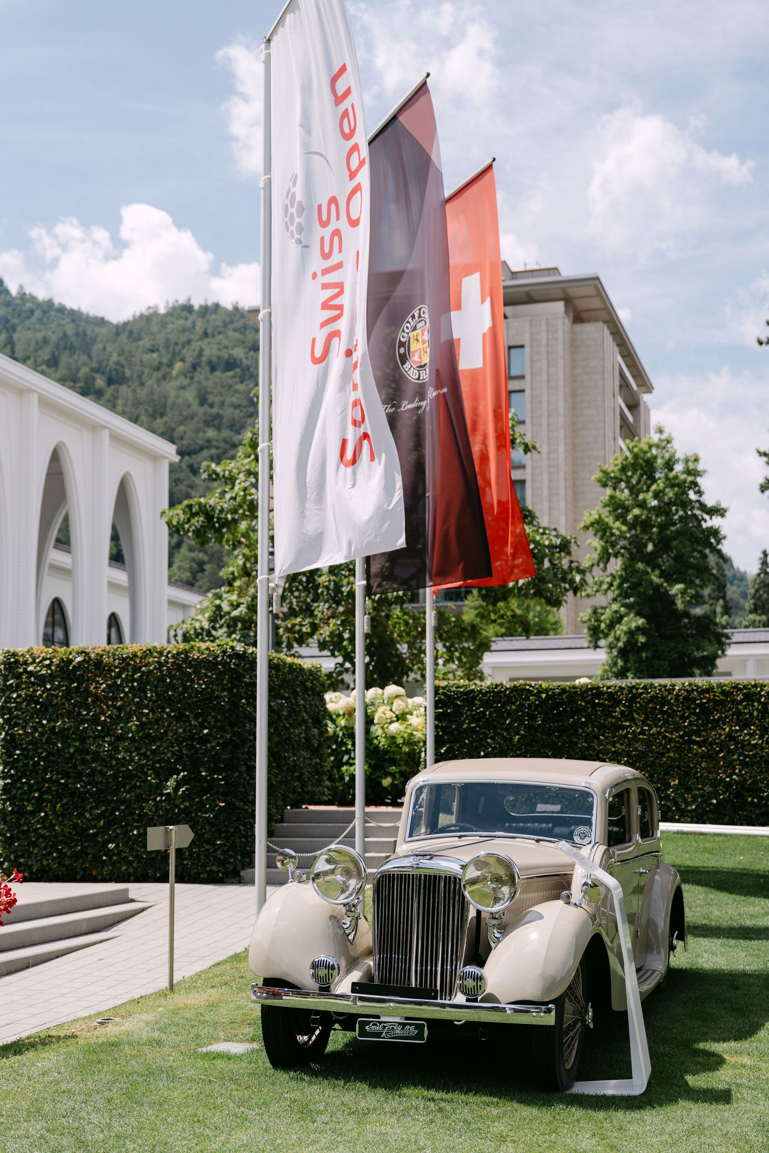 Classic vintage luxury cars displayed on grass with white flags and building facade in background.