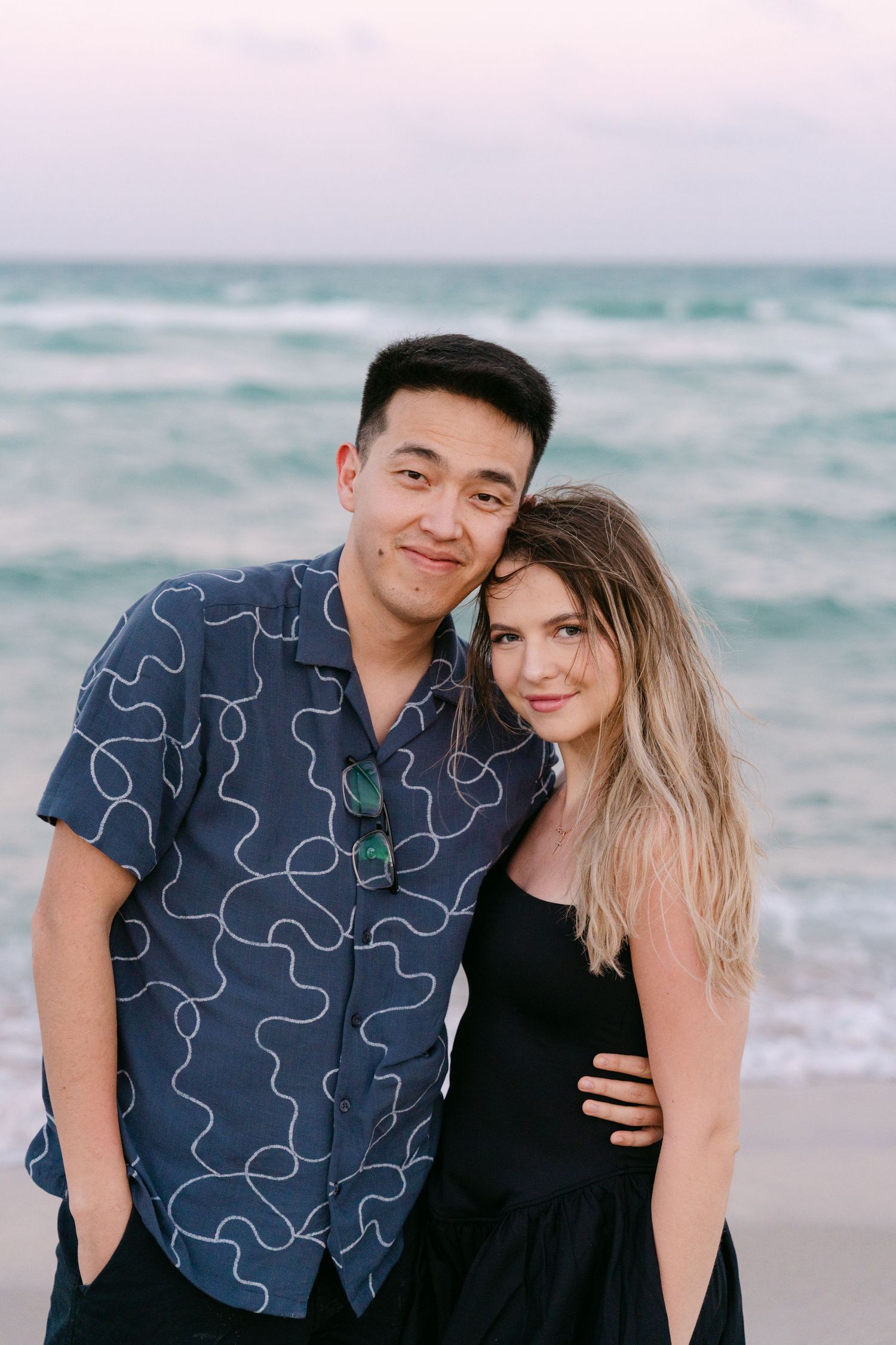 A couple poses casually together on the beach, with one wearing a patterned blue shirt.
