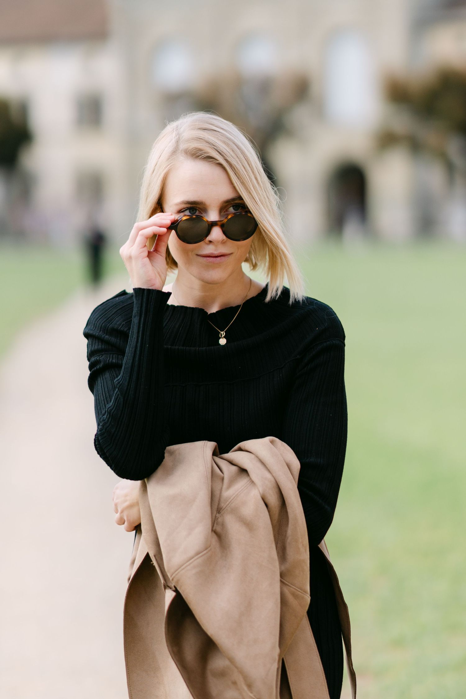 Woman adjusting sunglasses while wearing black sweater and holding beige coat outdoors.