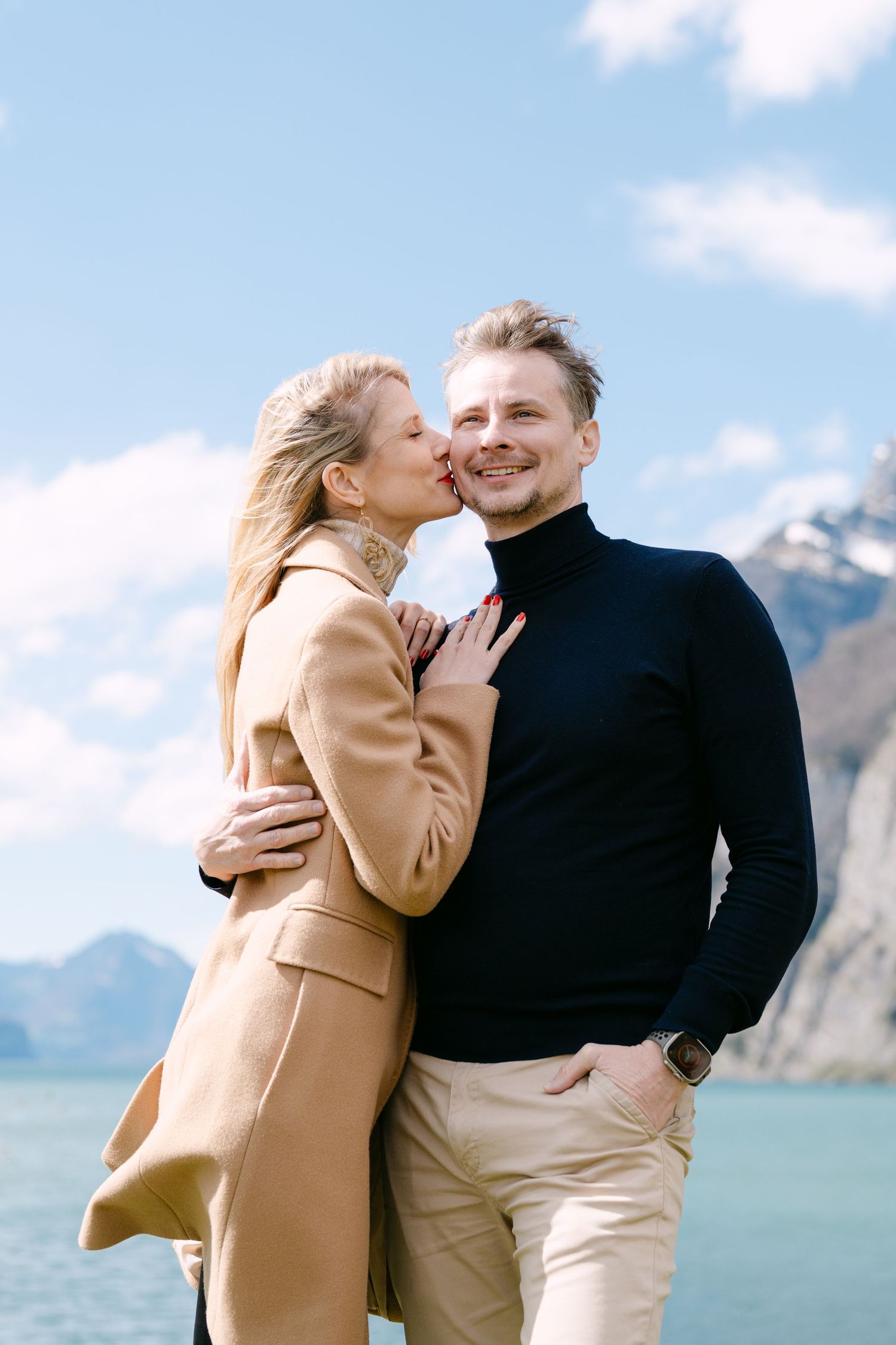 A romantic couple embraces against a mountain backdrop wearing a tan coat and black sweater.