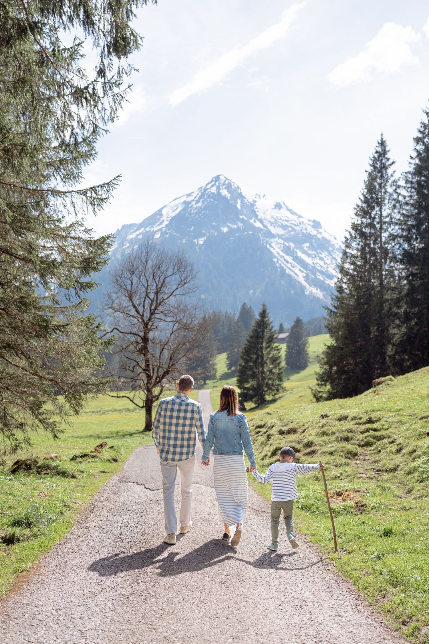 A family walks along a mountain trail with snow-capped Alps peaks in the background.