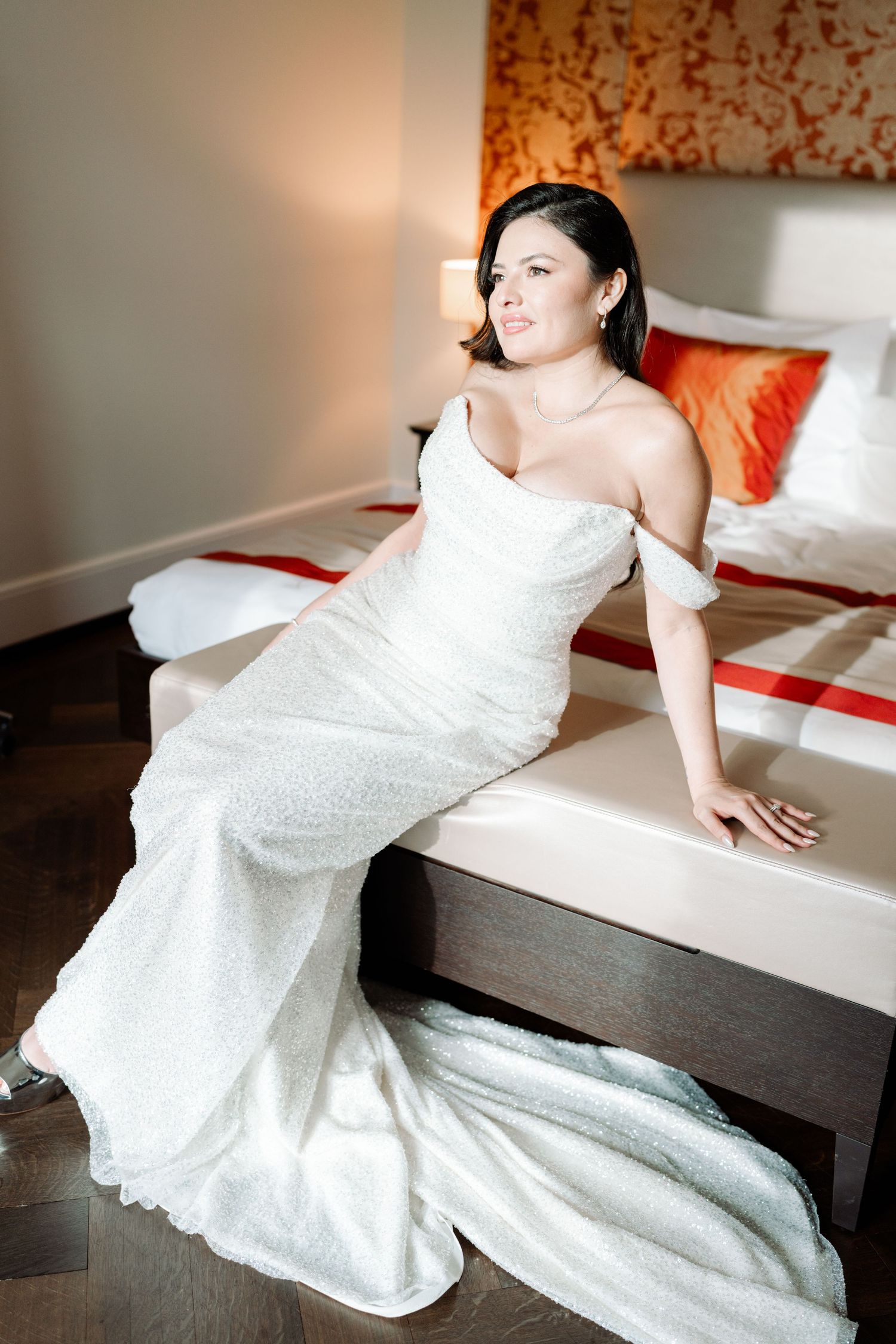Bride in fitted white gown poses elegantly on modern bed with orange accents.