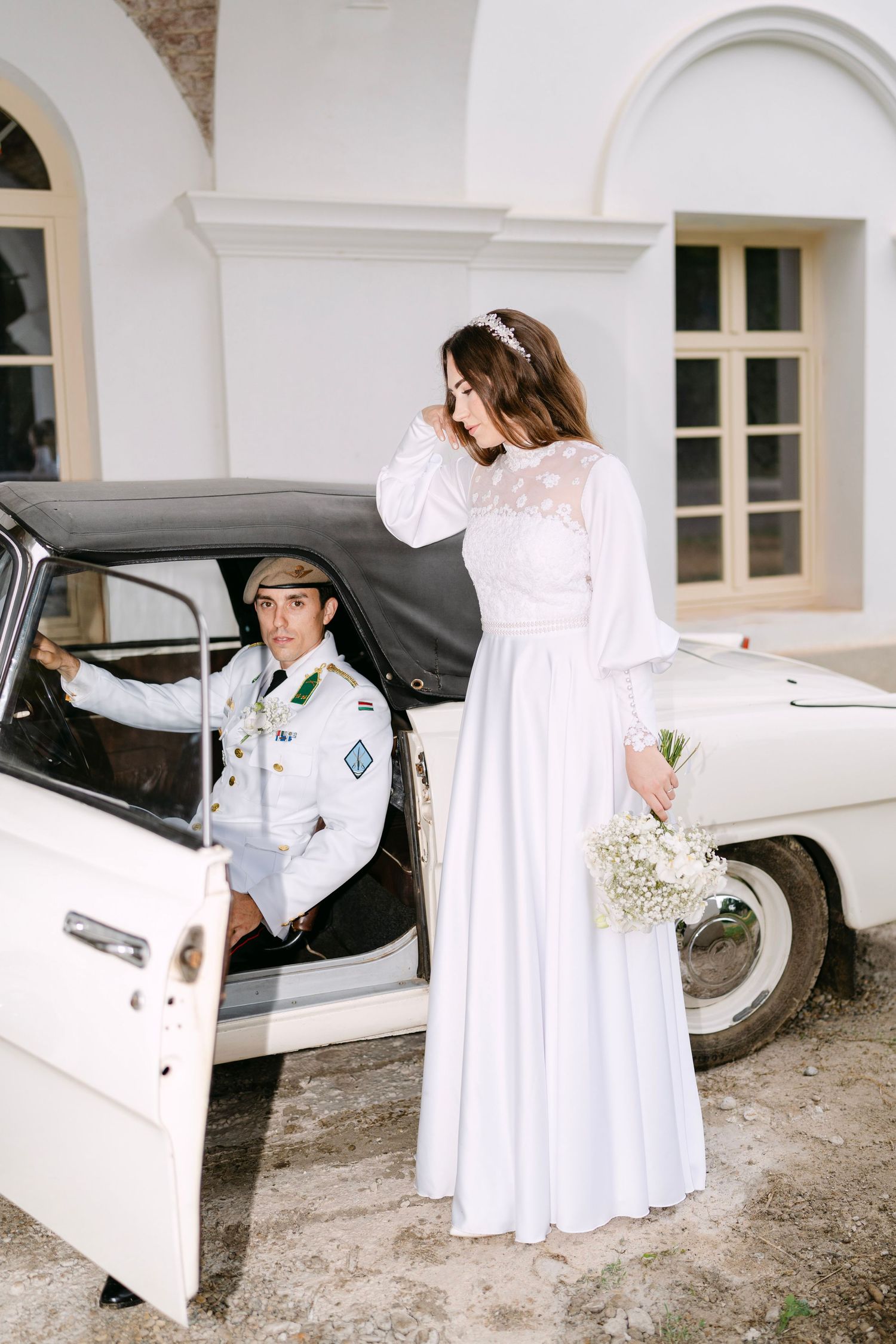 A couple in white attire poses near a vintage car under white archways.