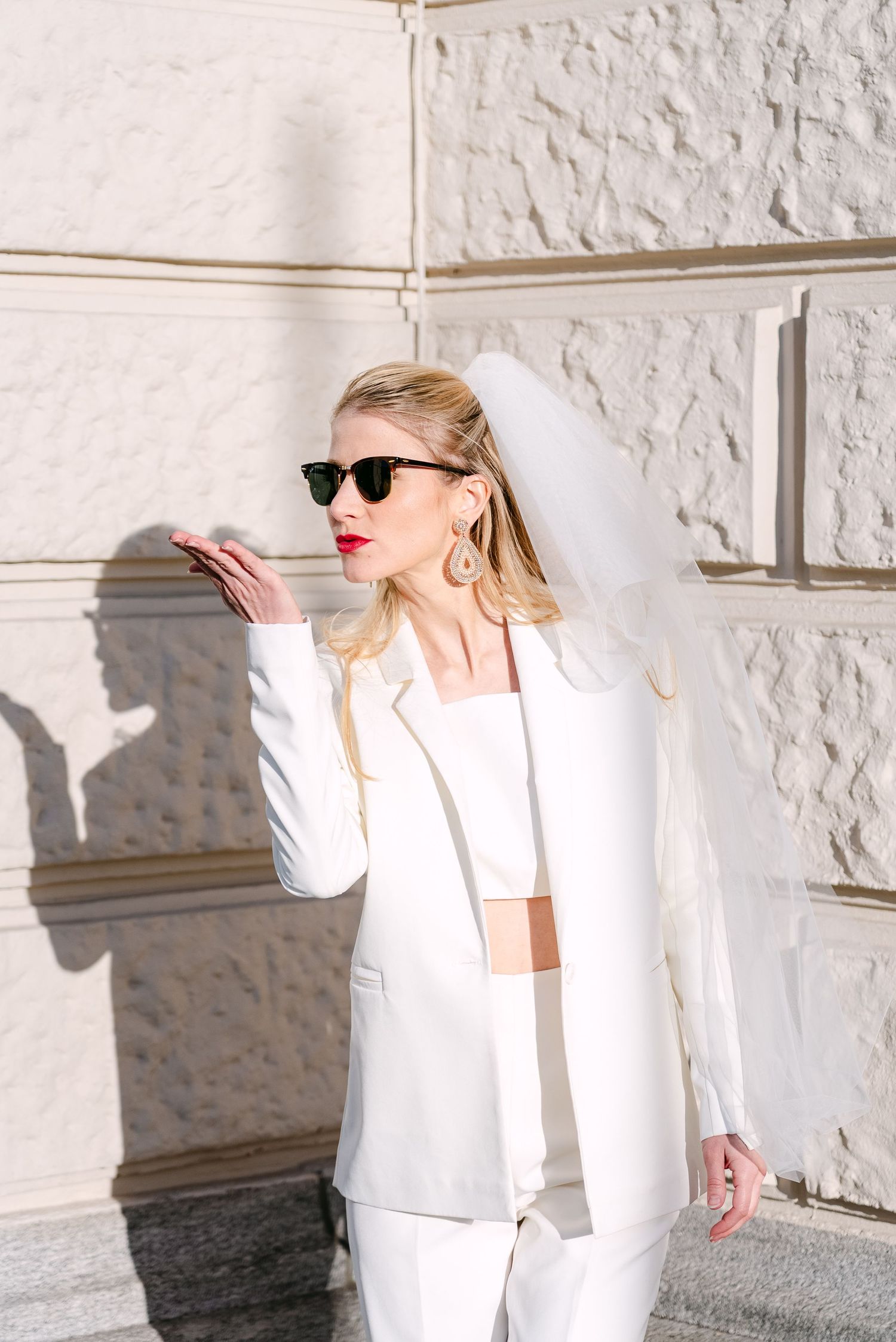 Woman in elegant white outfit poses dramatically against textured white wall.