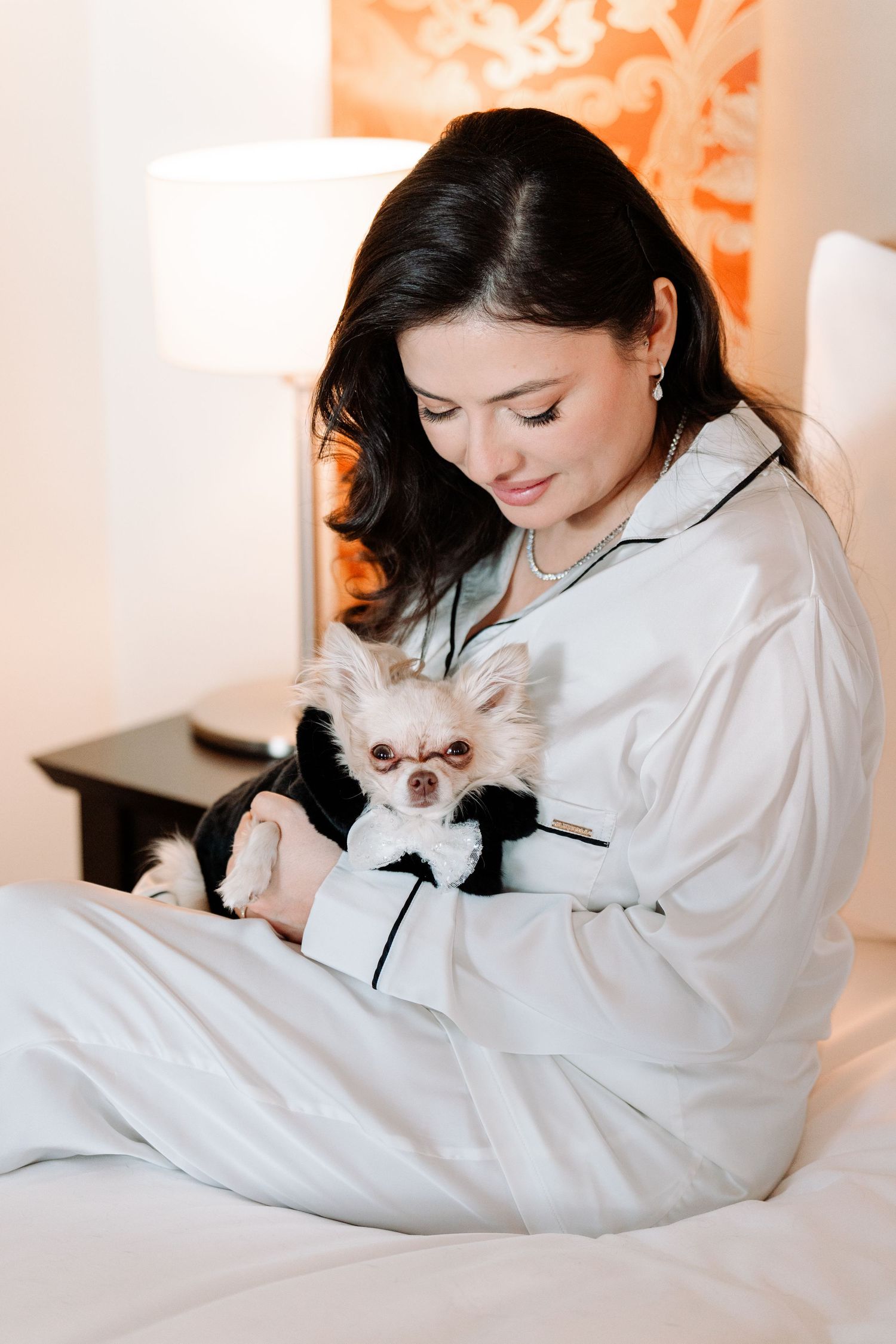 Woman in silk pajamas cuddles small white Chihuahua wearing bow tie on bed.