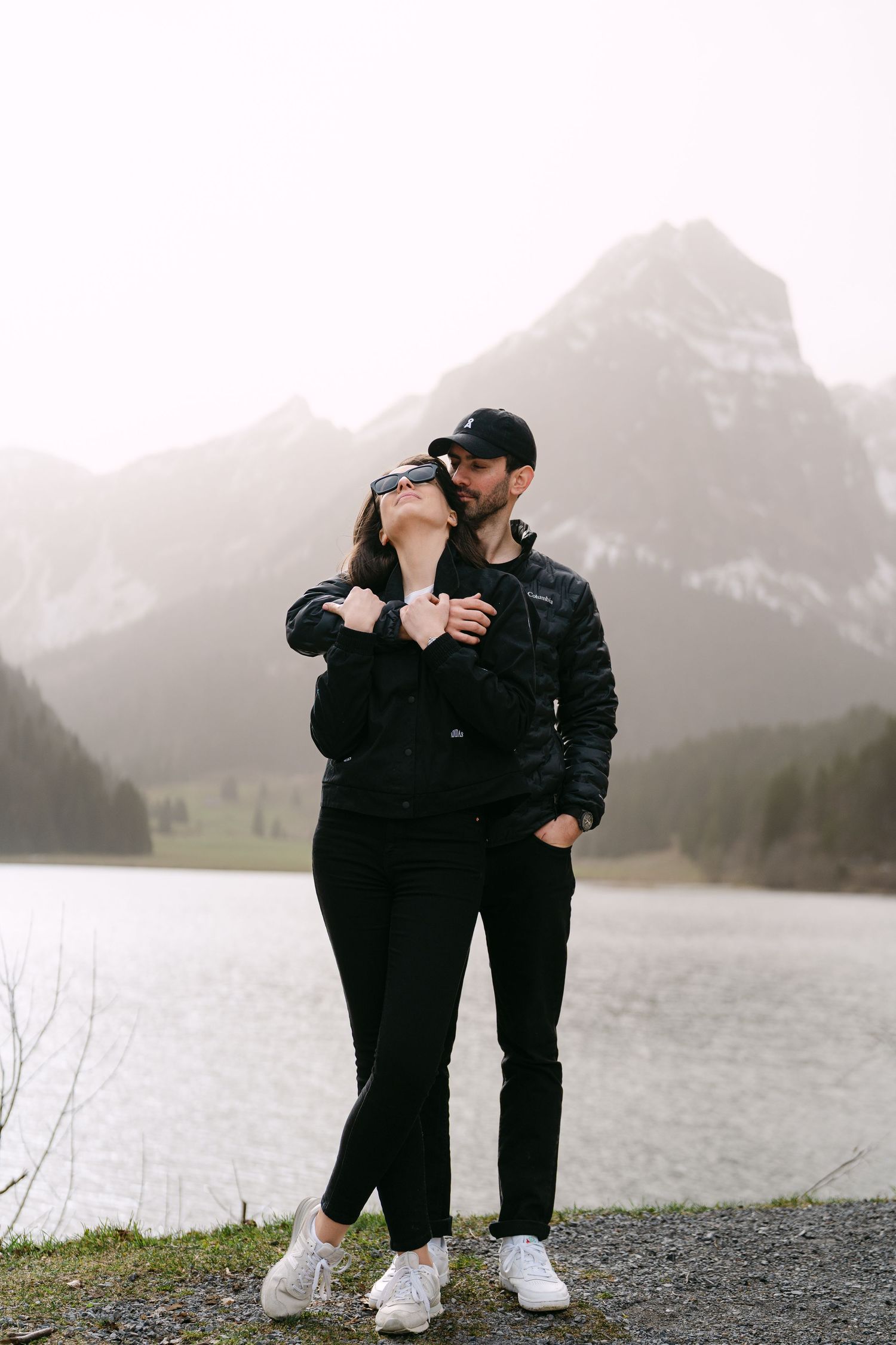 Couple in matching black outfits sharing tender moments by a mountain lake.