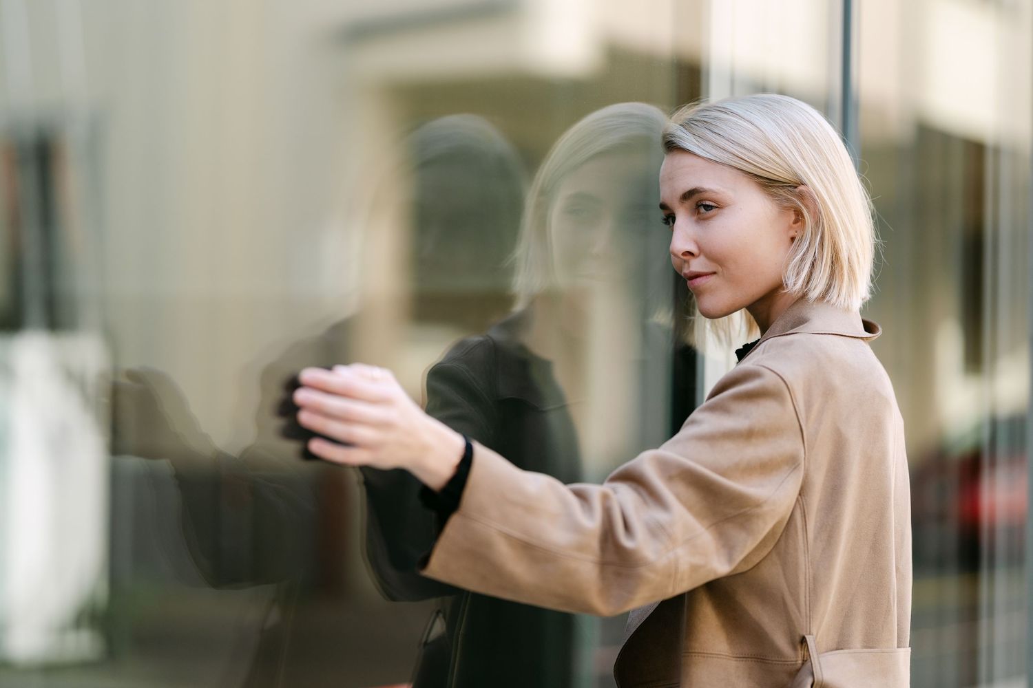 A person wearing a camel coat leans against reflective windows in an urban setting.