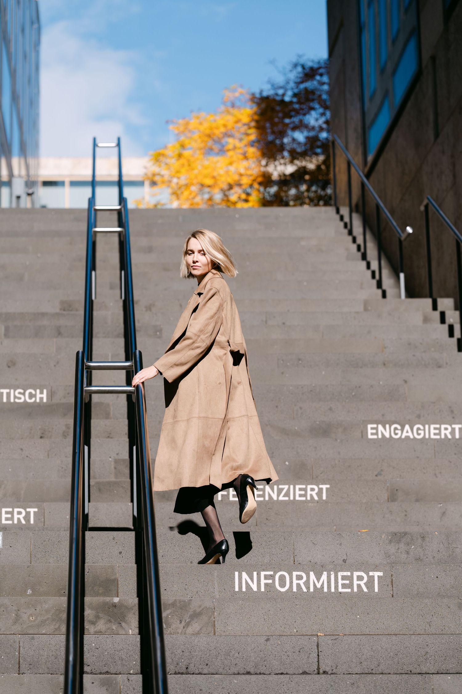 A person in a camel coat poses on concrete steps with autumn foliage in the background.