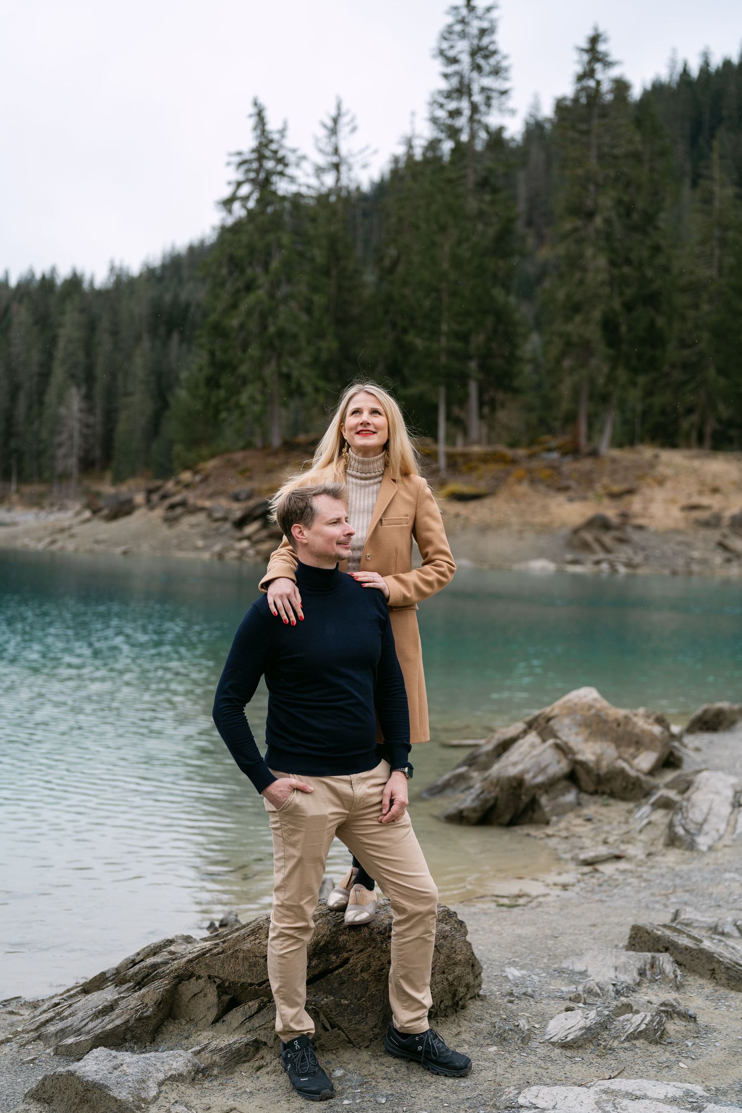 A couple stands on rocky shoreline with turquoise mountain lake and forest backdrop.