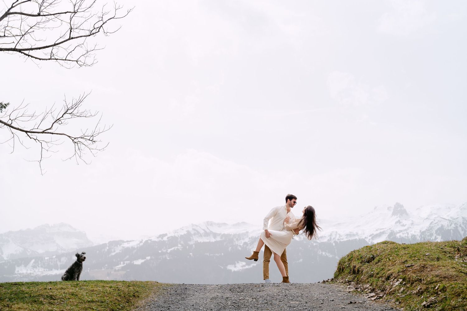 Two people and a dog share intimate moments on a hilltop with snow-capped mountains in view.