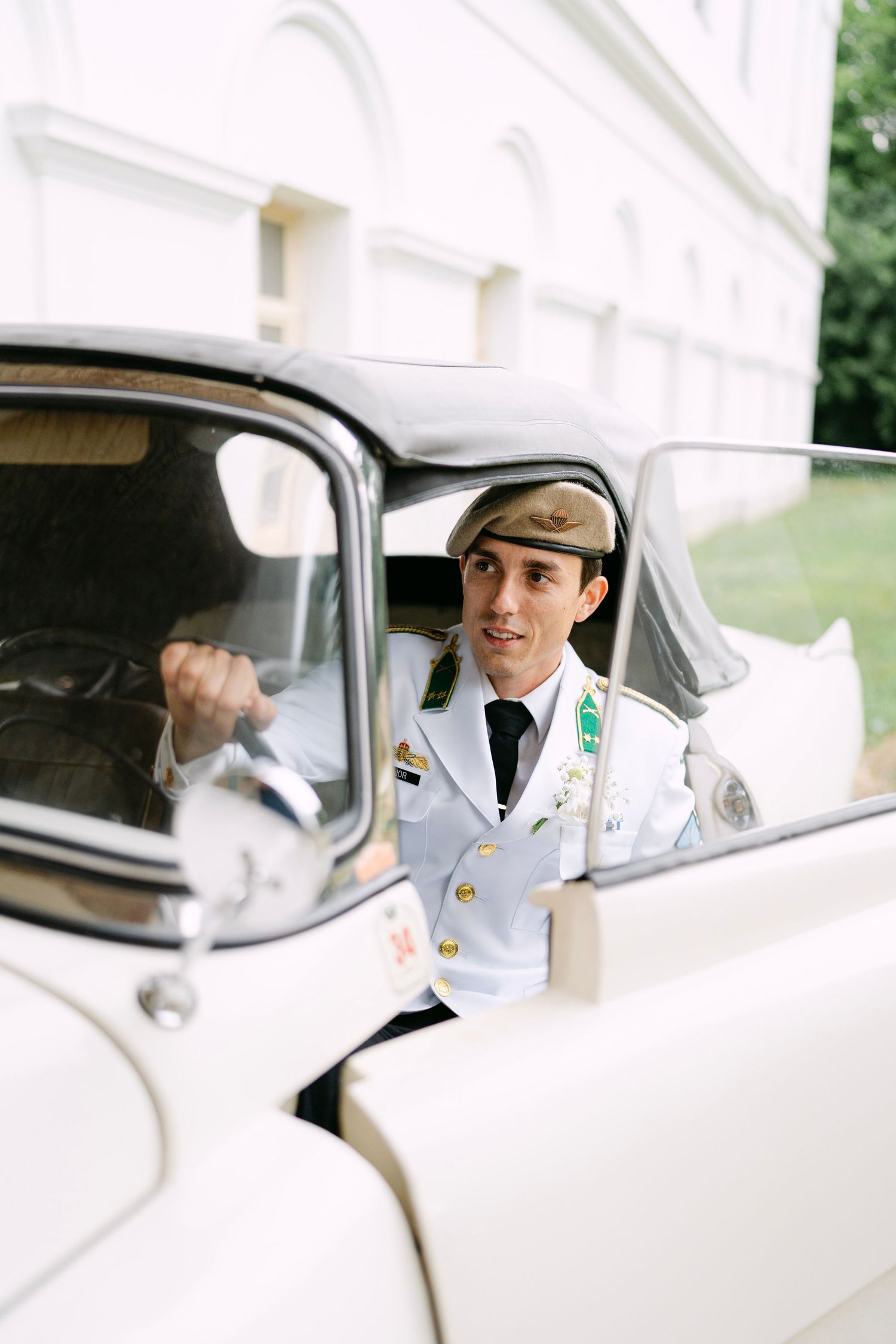 A classic white vintage car with a driver visible through the window on a sunny day.