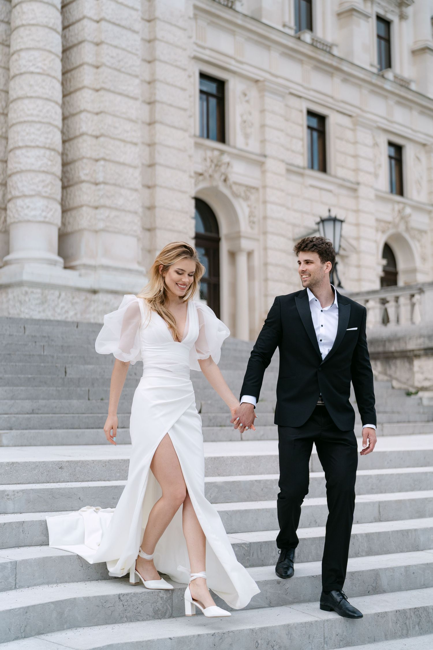 Playful wedding couple descends grand marble staircase in formal attire.