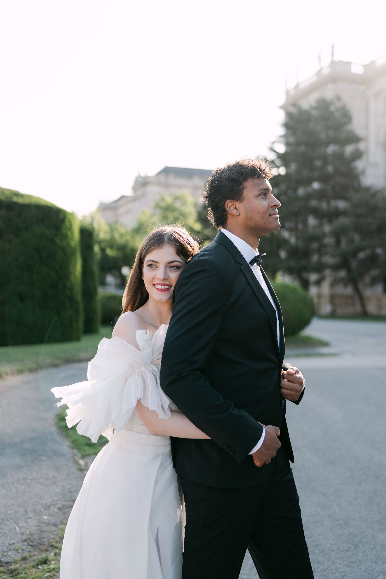 Playful wedding couple poses together on path near ornate building.