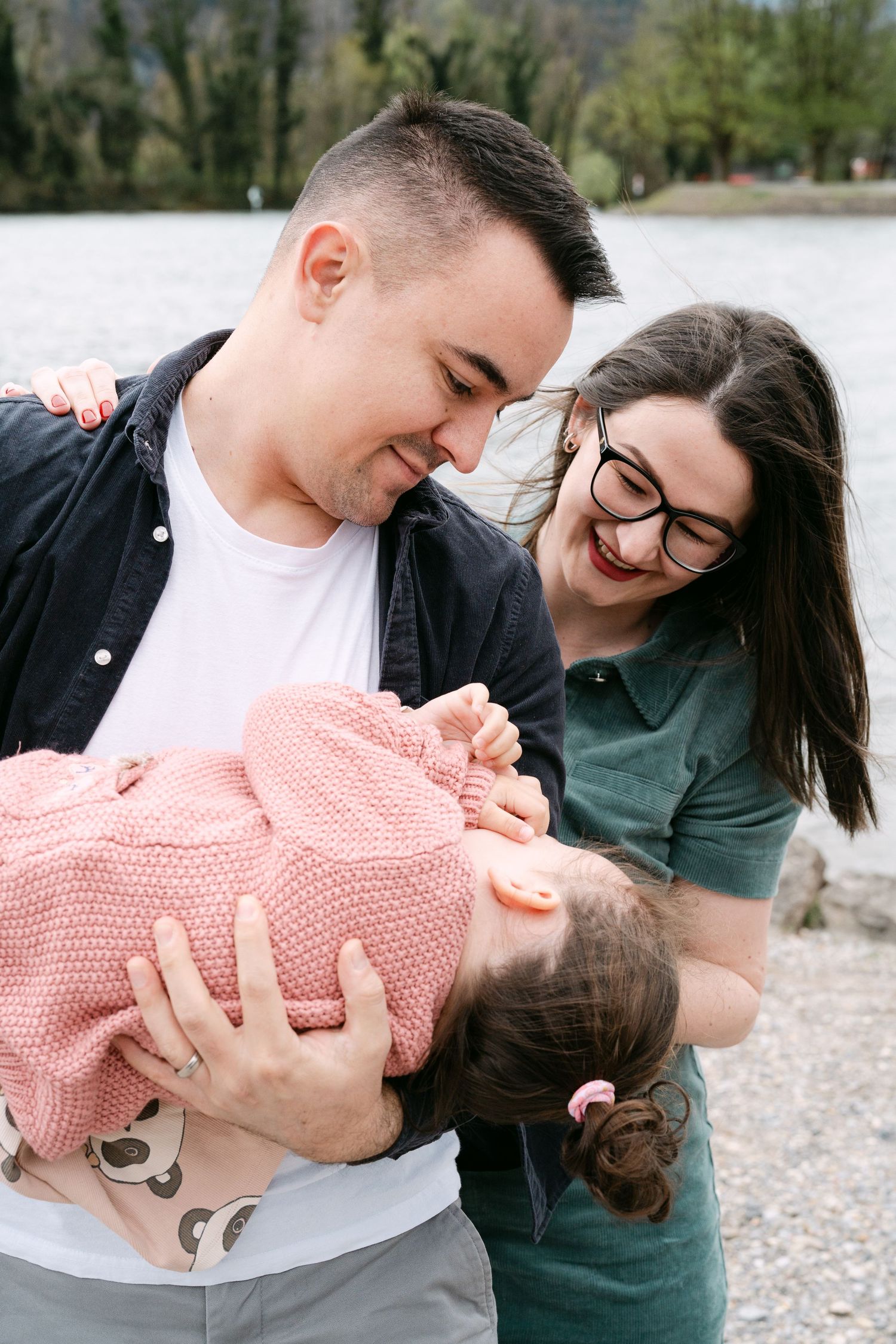 A couple shares a joyful moment with their baby wearing a pink knit sweater by the water.