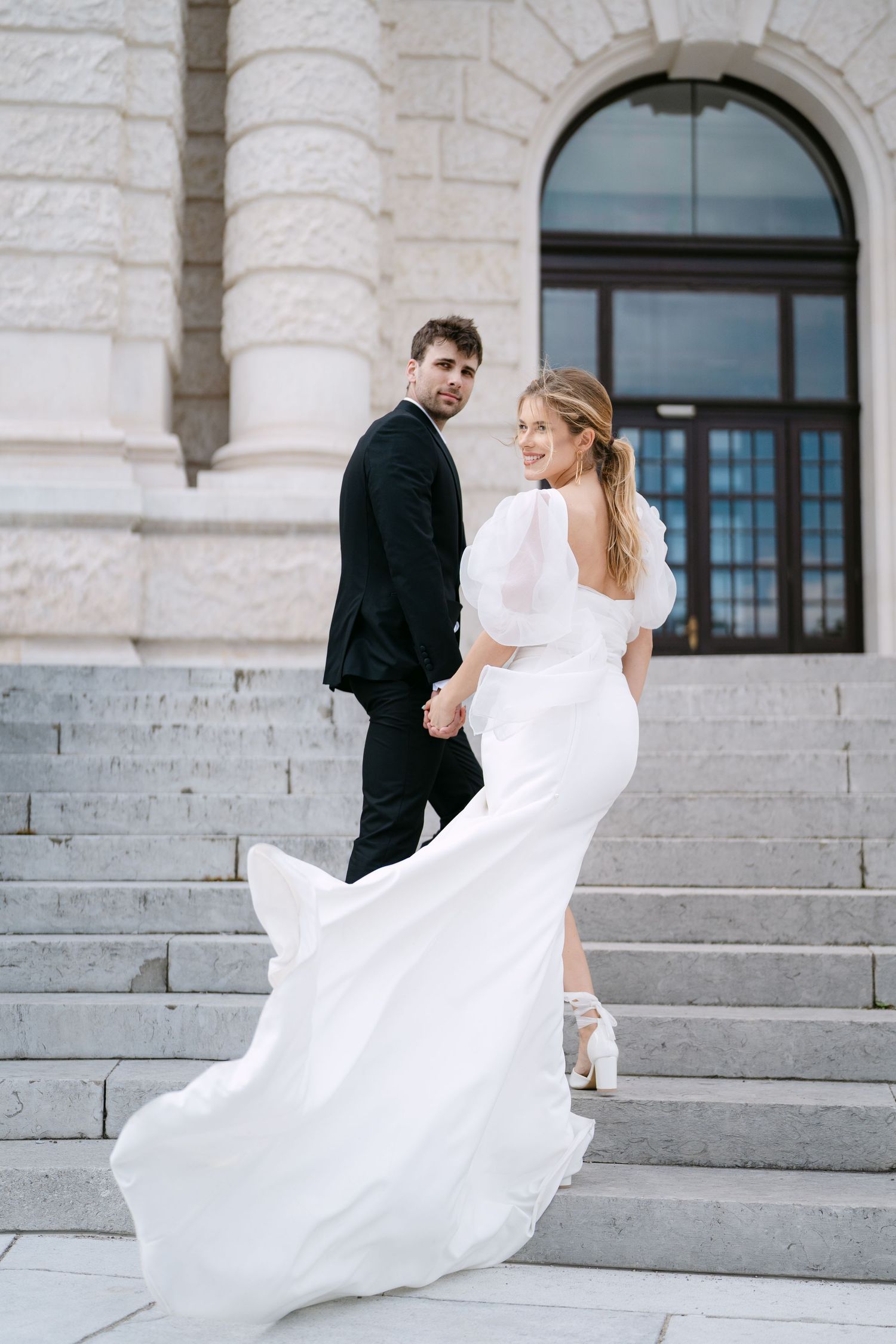 Elegant couple poses on stone steps of historic building during wedding photoshoot.