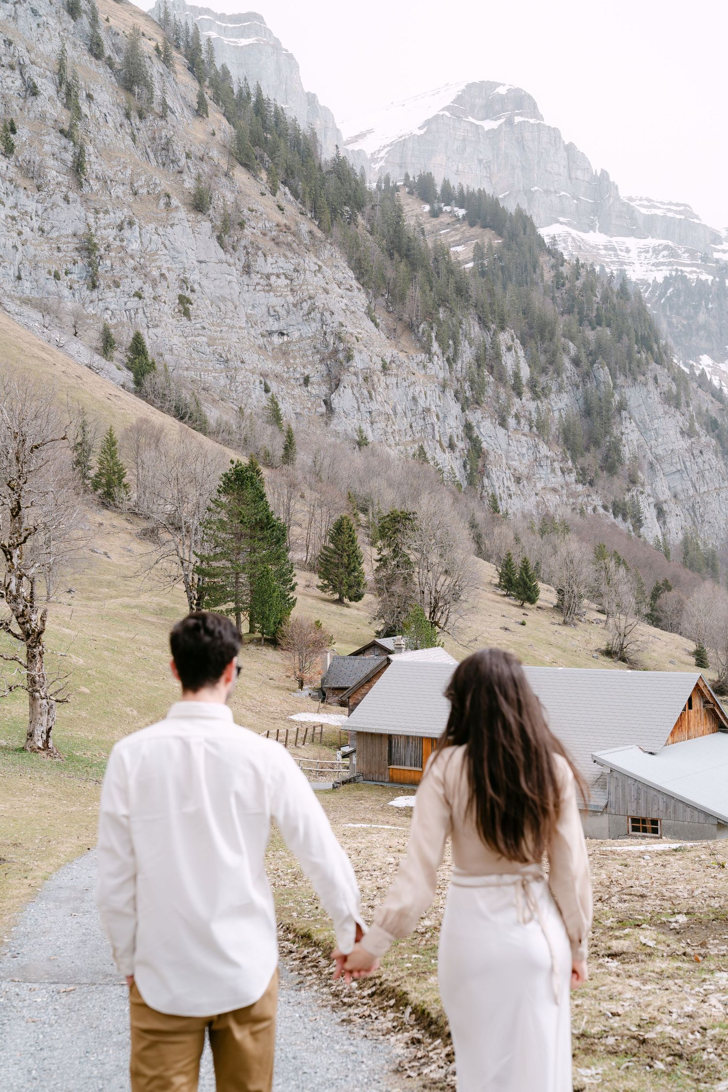 Couple walking hand in hand on mountain path with rustic cabin and alpine scenery.
