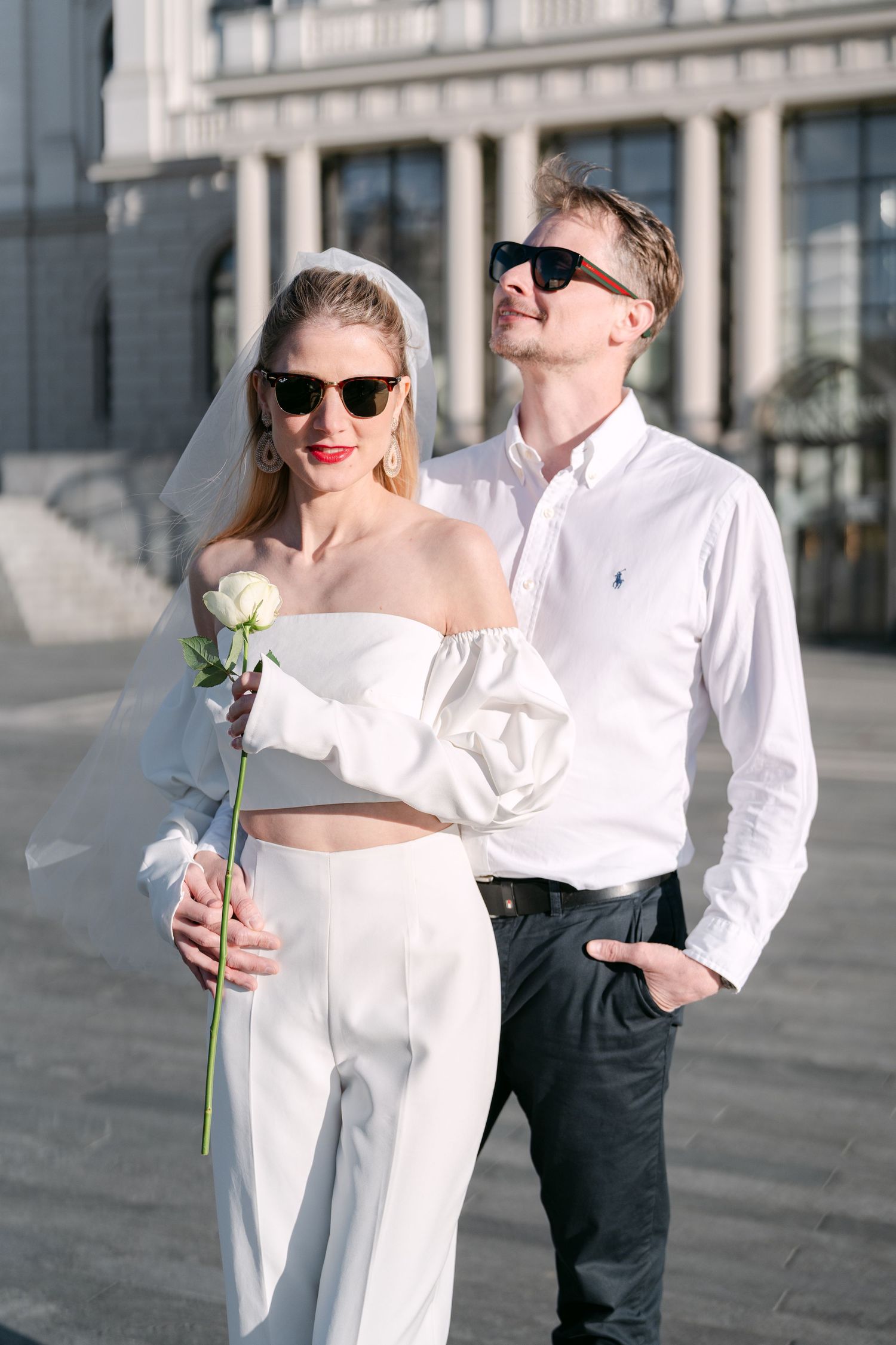 Contemporary wedding portraits with white rose and sunglasses against city architecture.