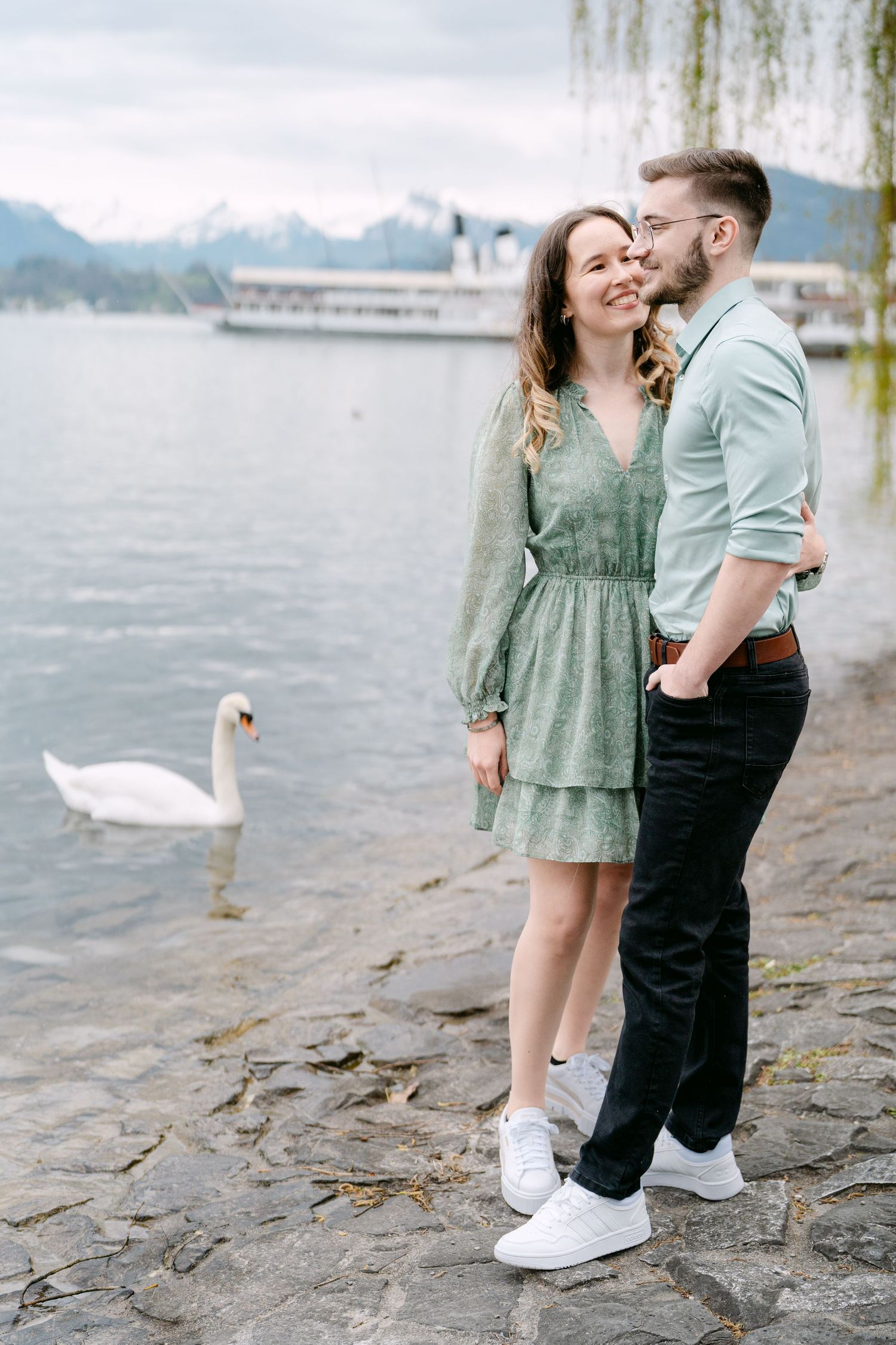A couple in green and black outfits poses by a lake with a swan in the background.