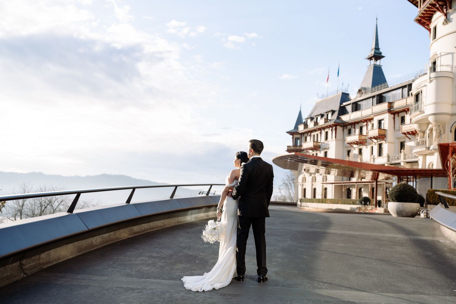 Couple in wedding attire stands on scenic European waterfront terrace.