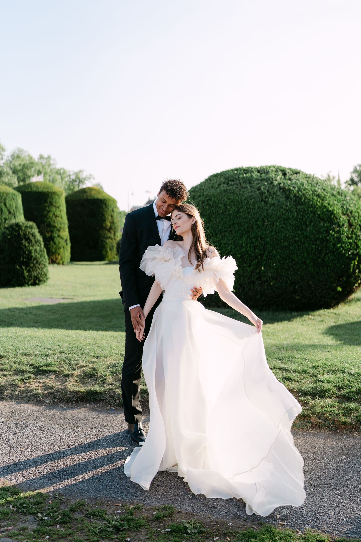 Elegant wedding dress flows in the breeze as couple poses near garden hedges.