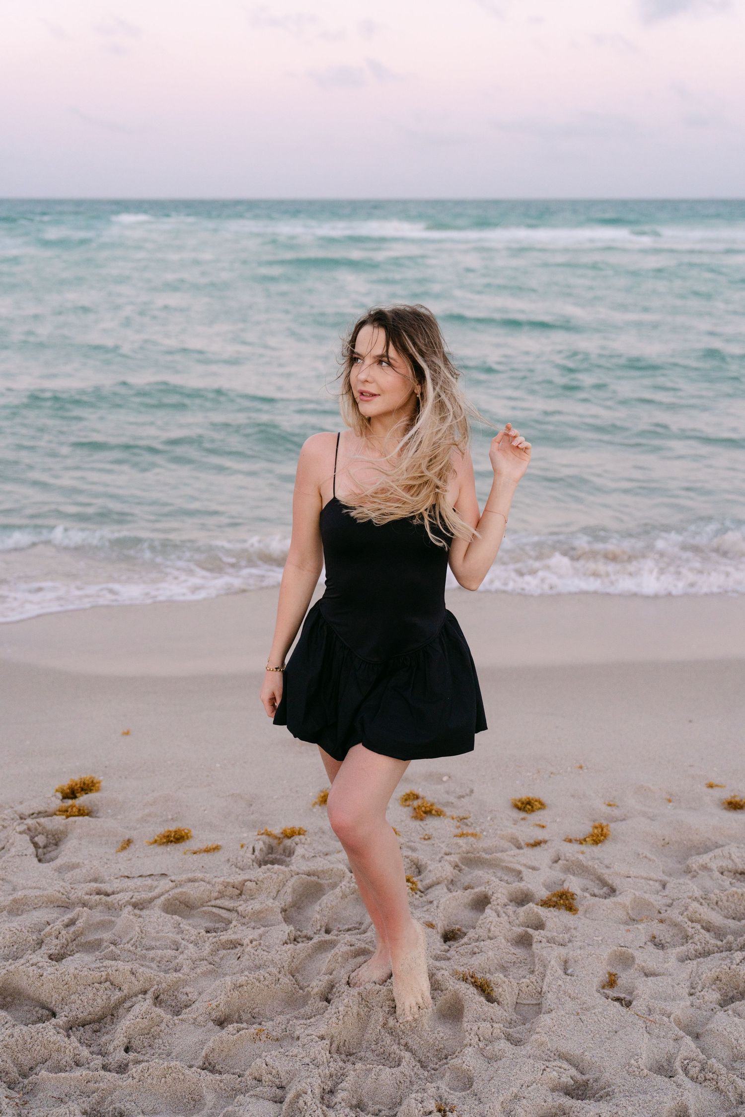 Woman in black dress poses playfully on sandy beach at sunset.