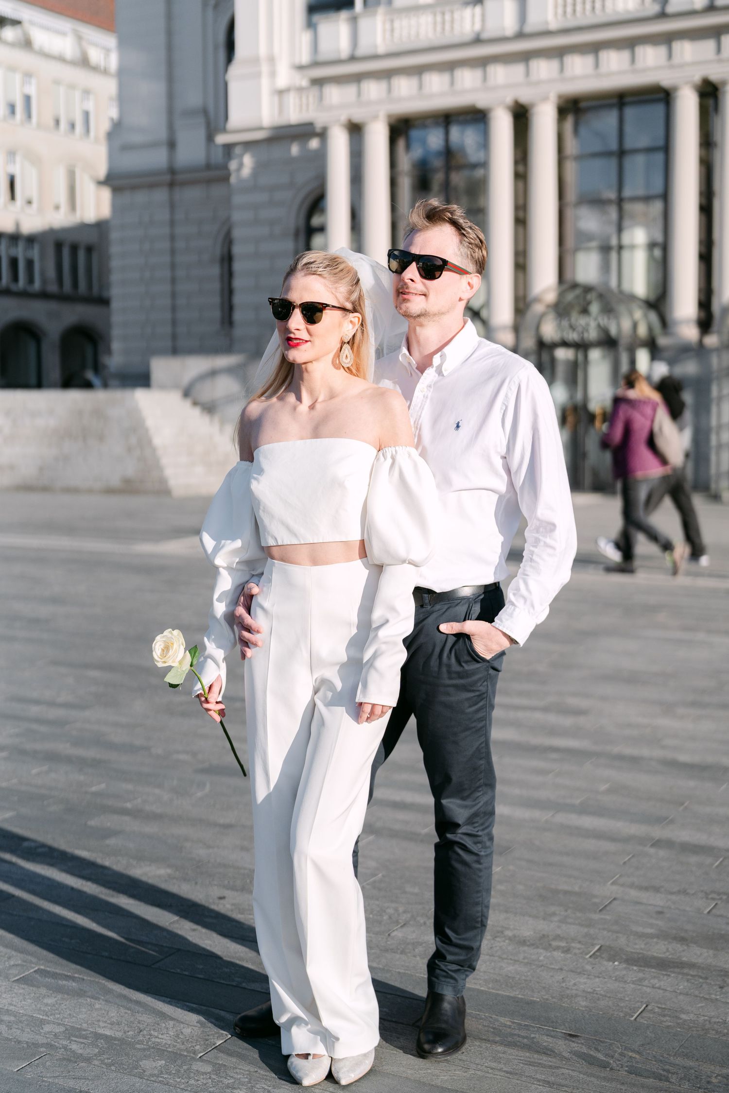 Stylish couple in white and black attire posing in urban square with sunglasses.