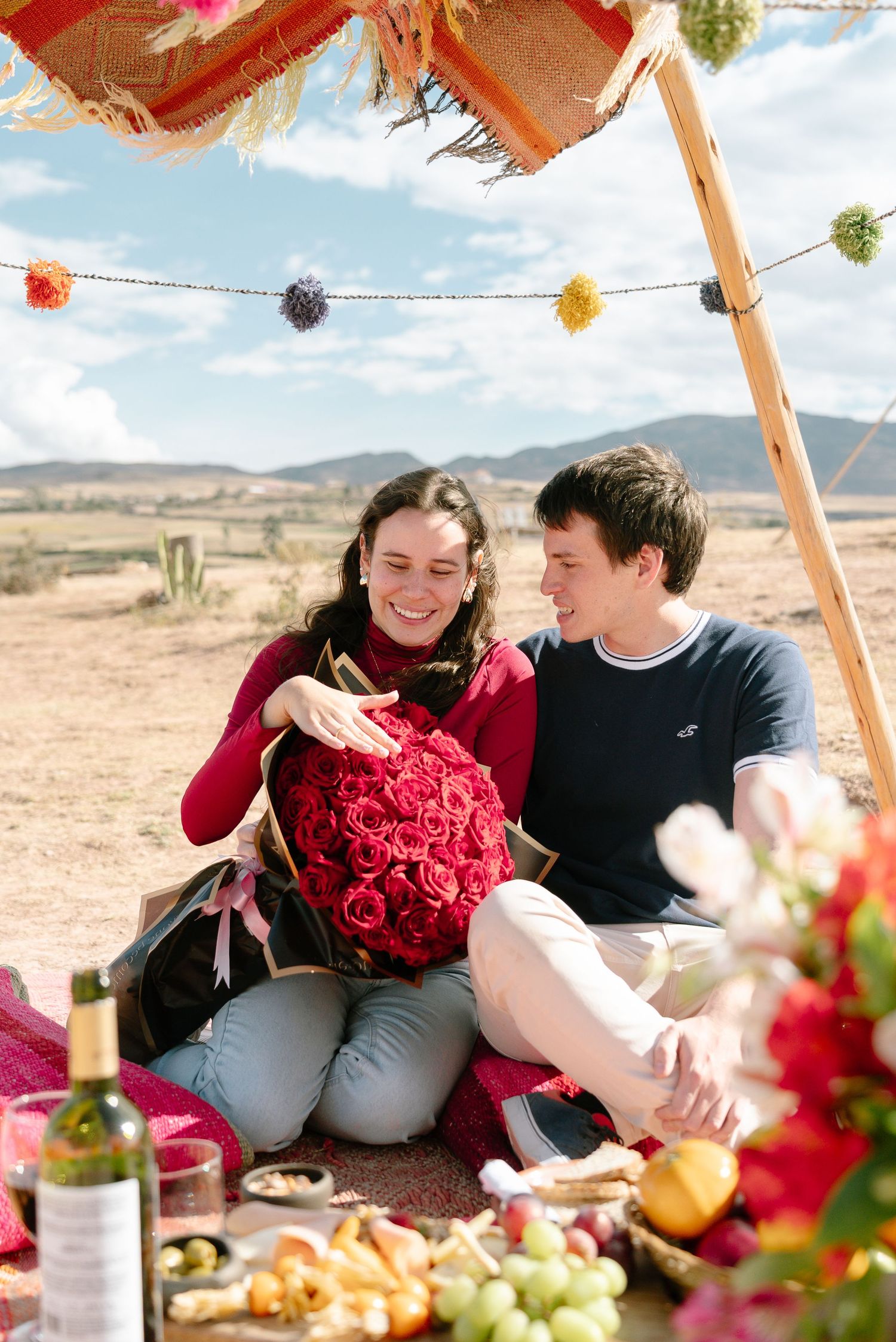 Romantic picnic setup with red roses and food spread under decorated canopy in desert setting.