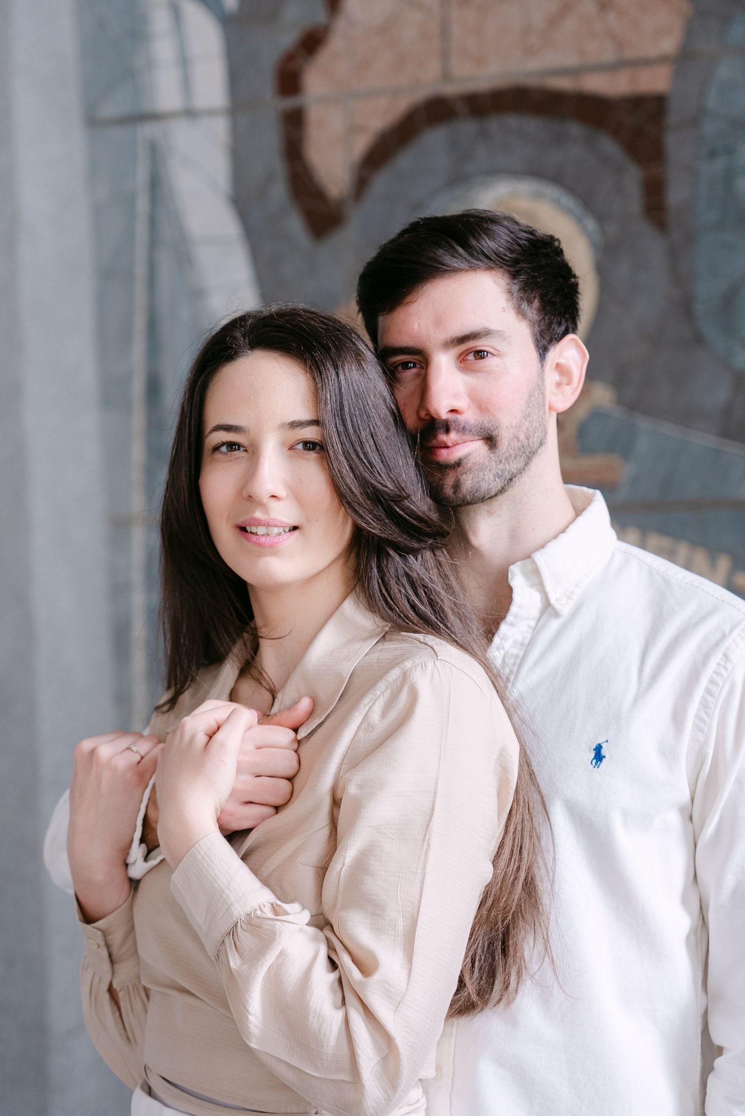 Romantic indoor photoshoot with couple in light colored outfits against brick wall background.