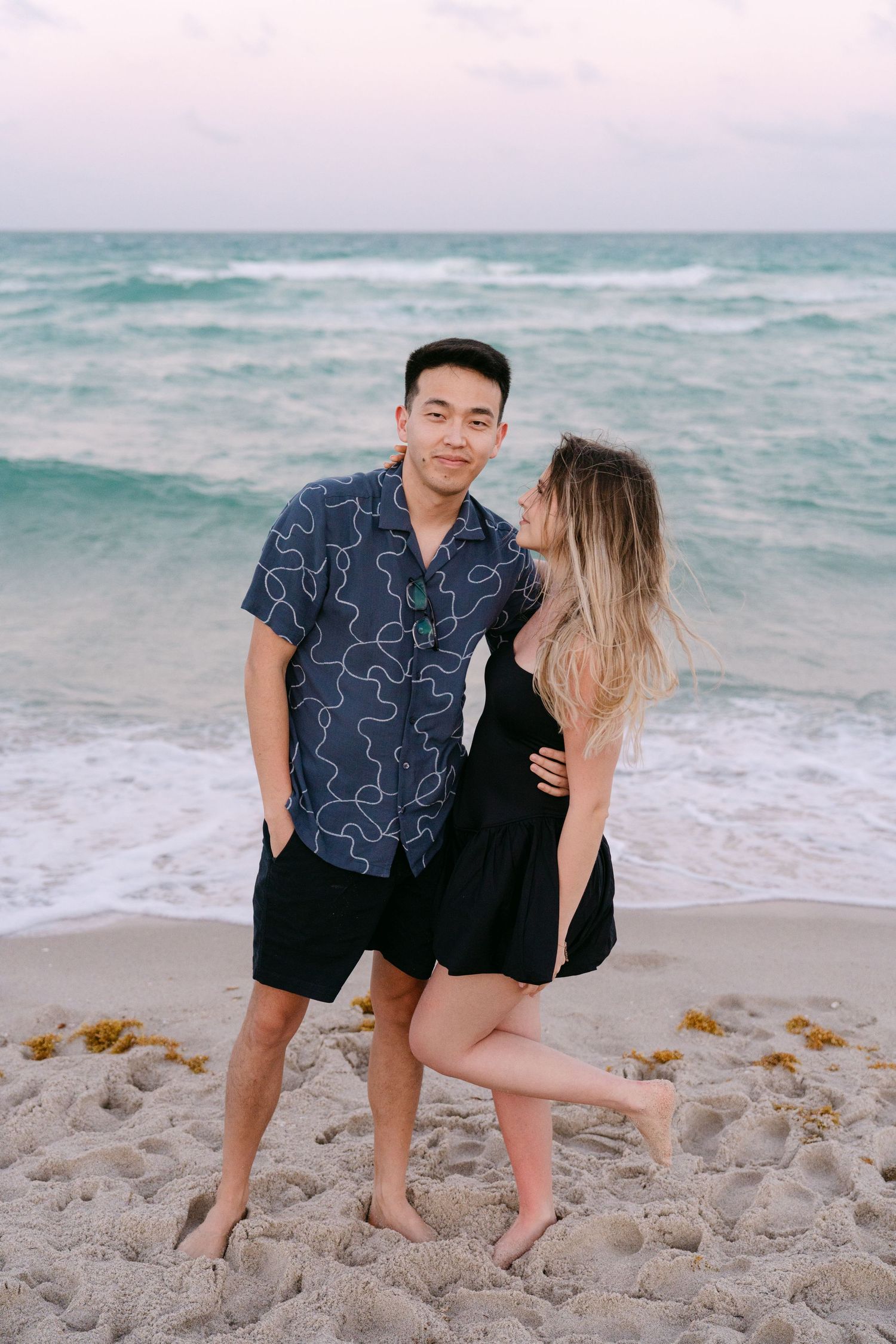 Two people playfully interact on the beach sand with ocean waves behind them.