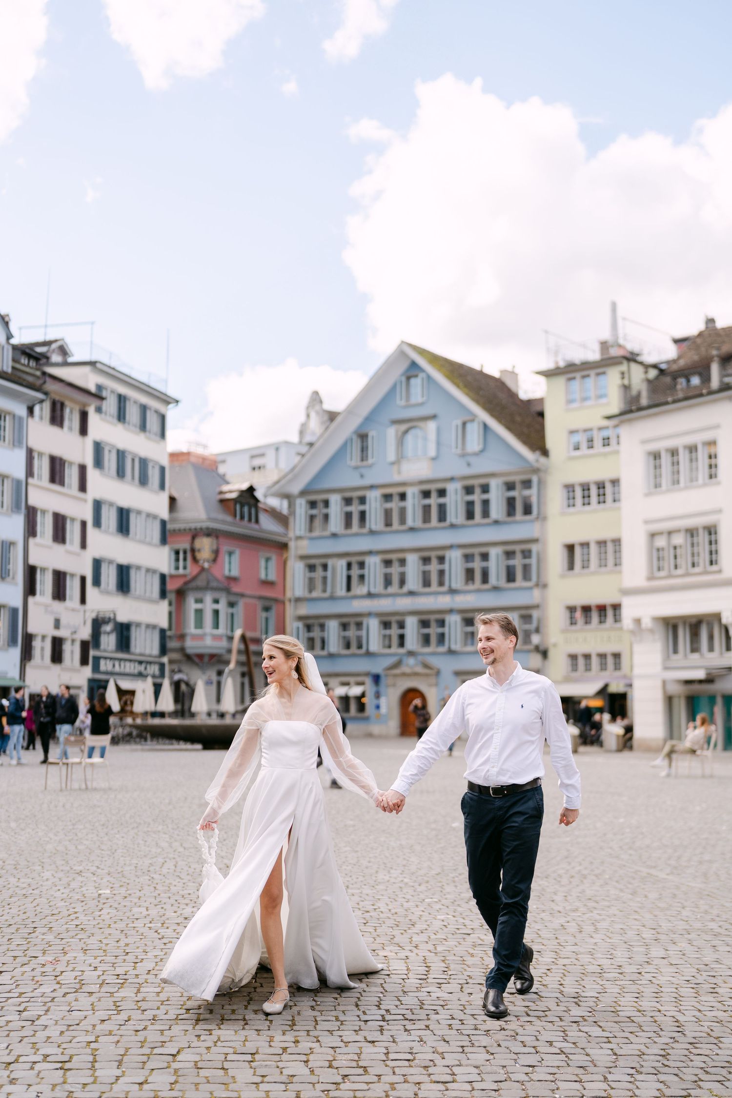 Newlyweds dance playfully in cobblestone town square surrounded by historic buildings.
