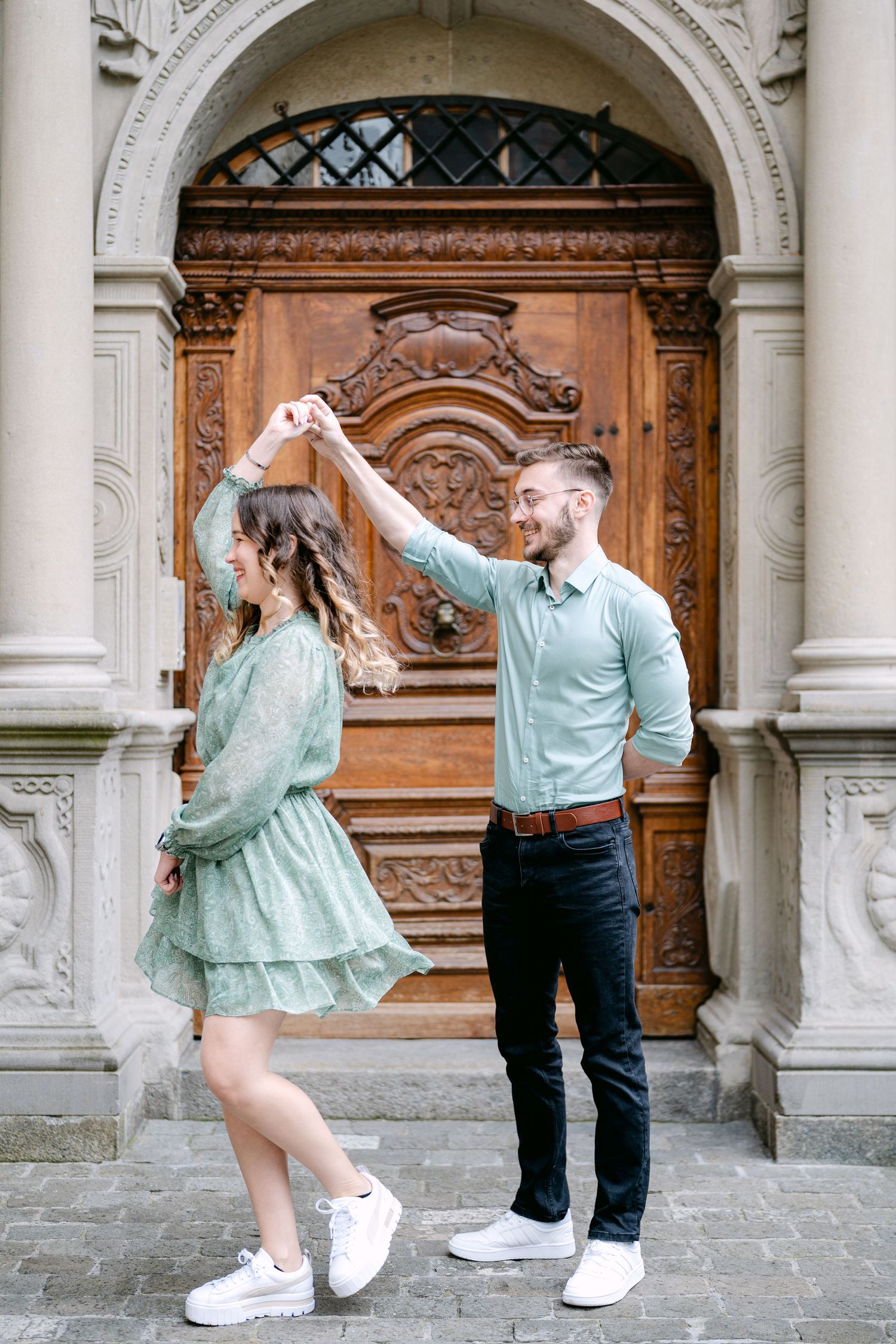 A couple dances playfully in front of a historic stone archway wearing matching sneakers.