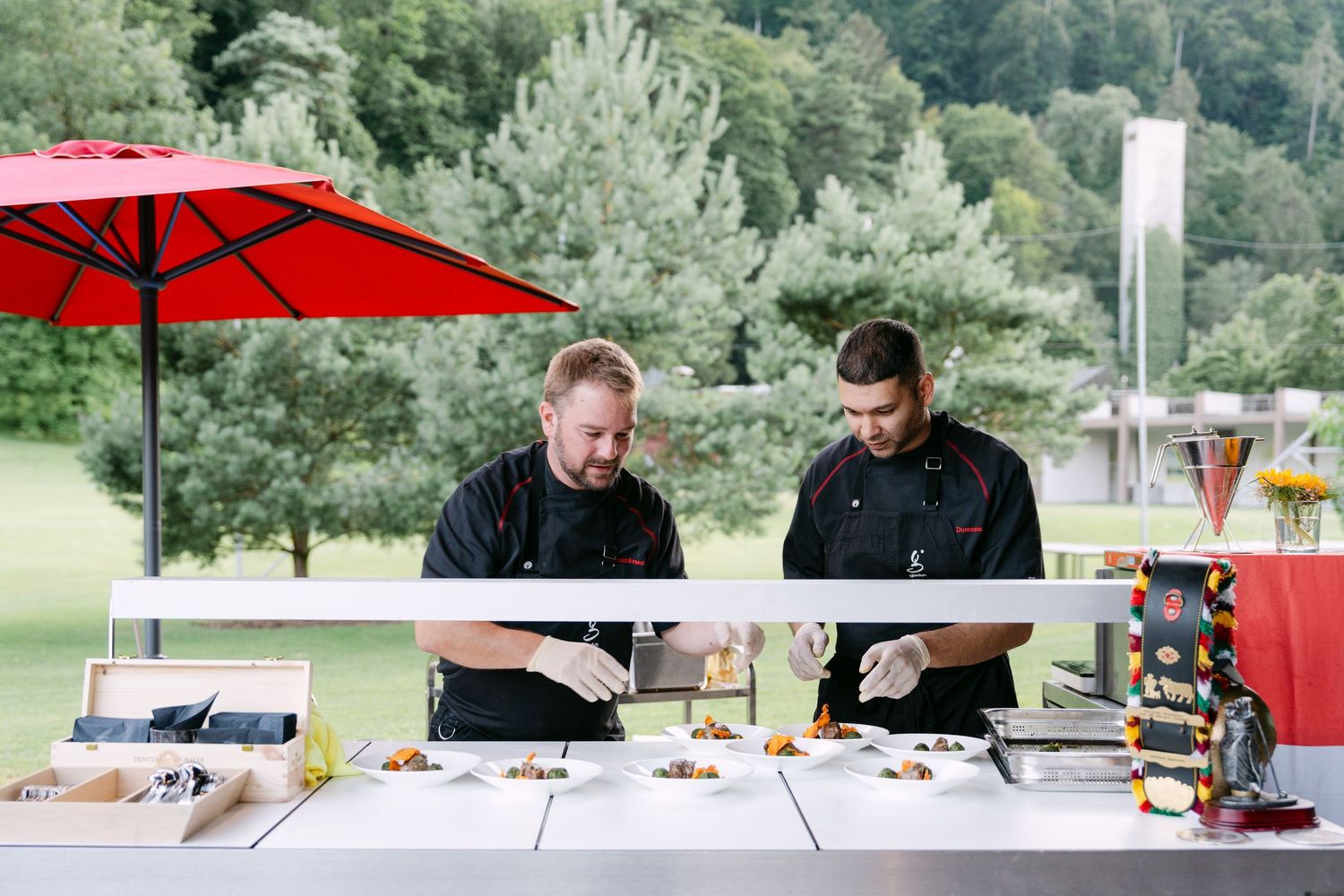 Chefs prepare food at an outdoor catering station with a red umbrella.
