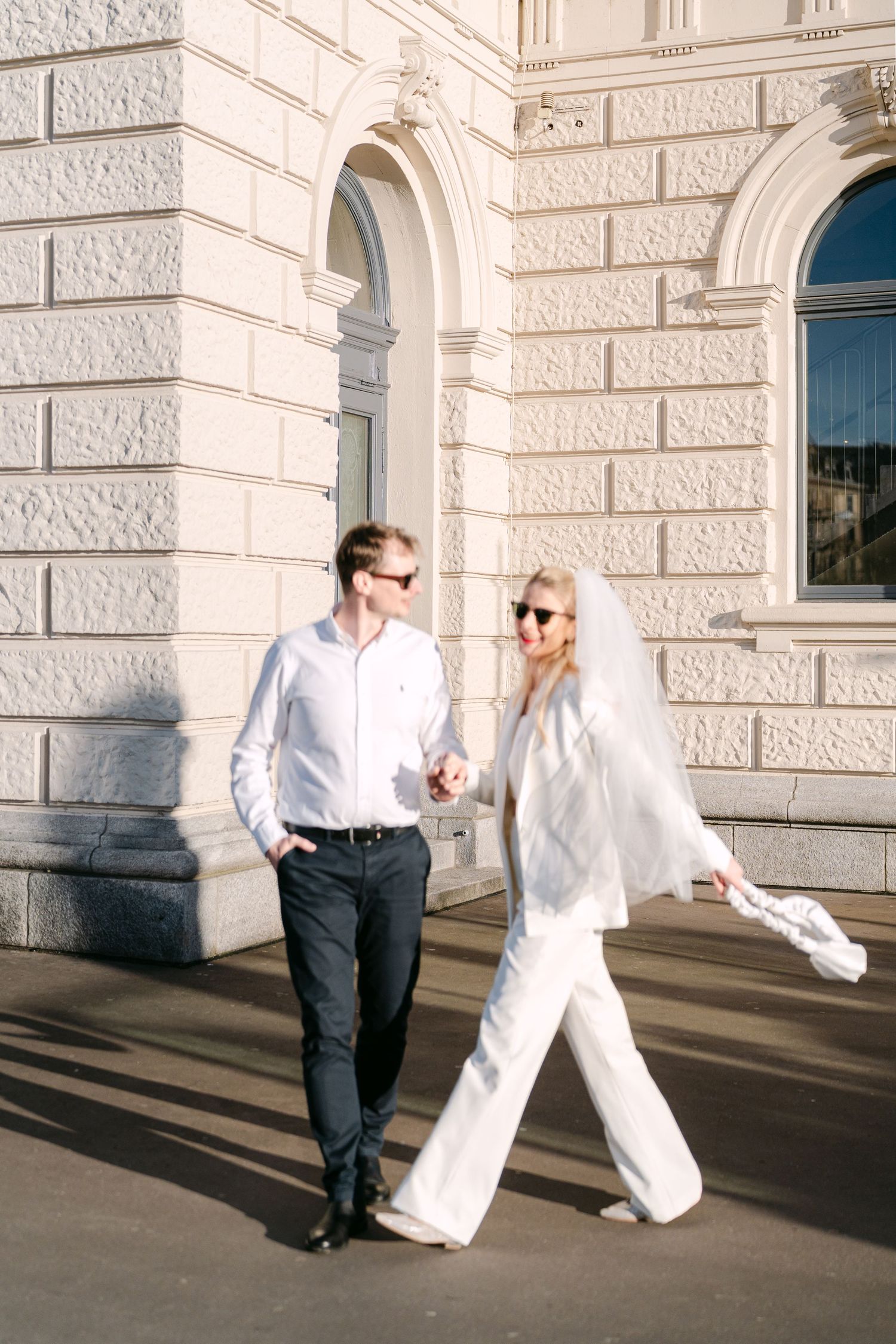 Couple in contrasting black and white outfits dance joyfully along stone building wall.