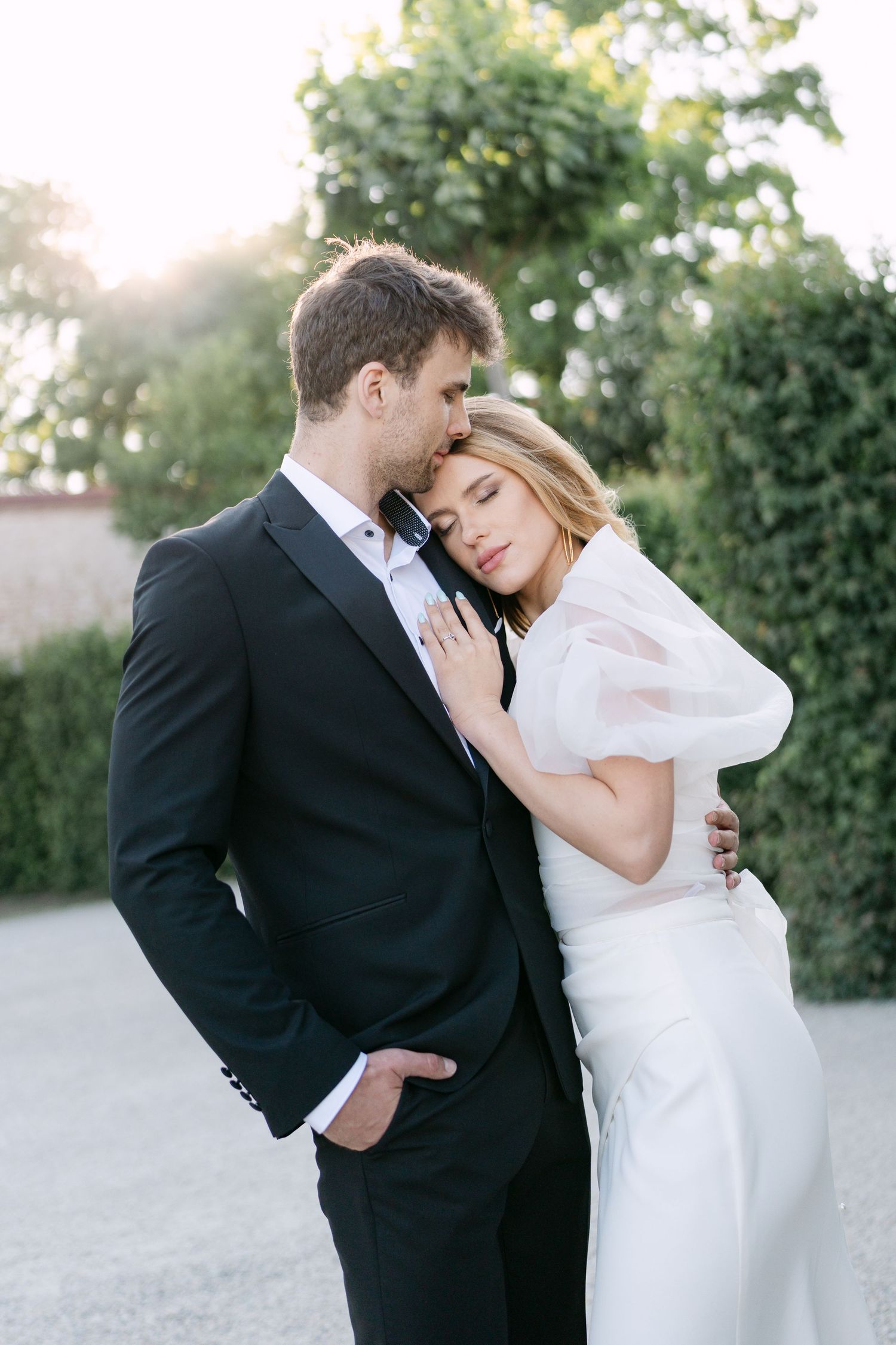 A romantic wedding portrait of a couple embracing outdoors at sunset with lush greenery in the background.