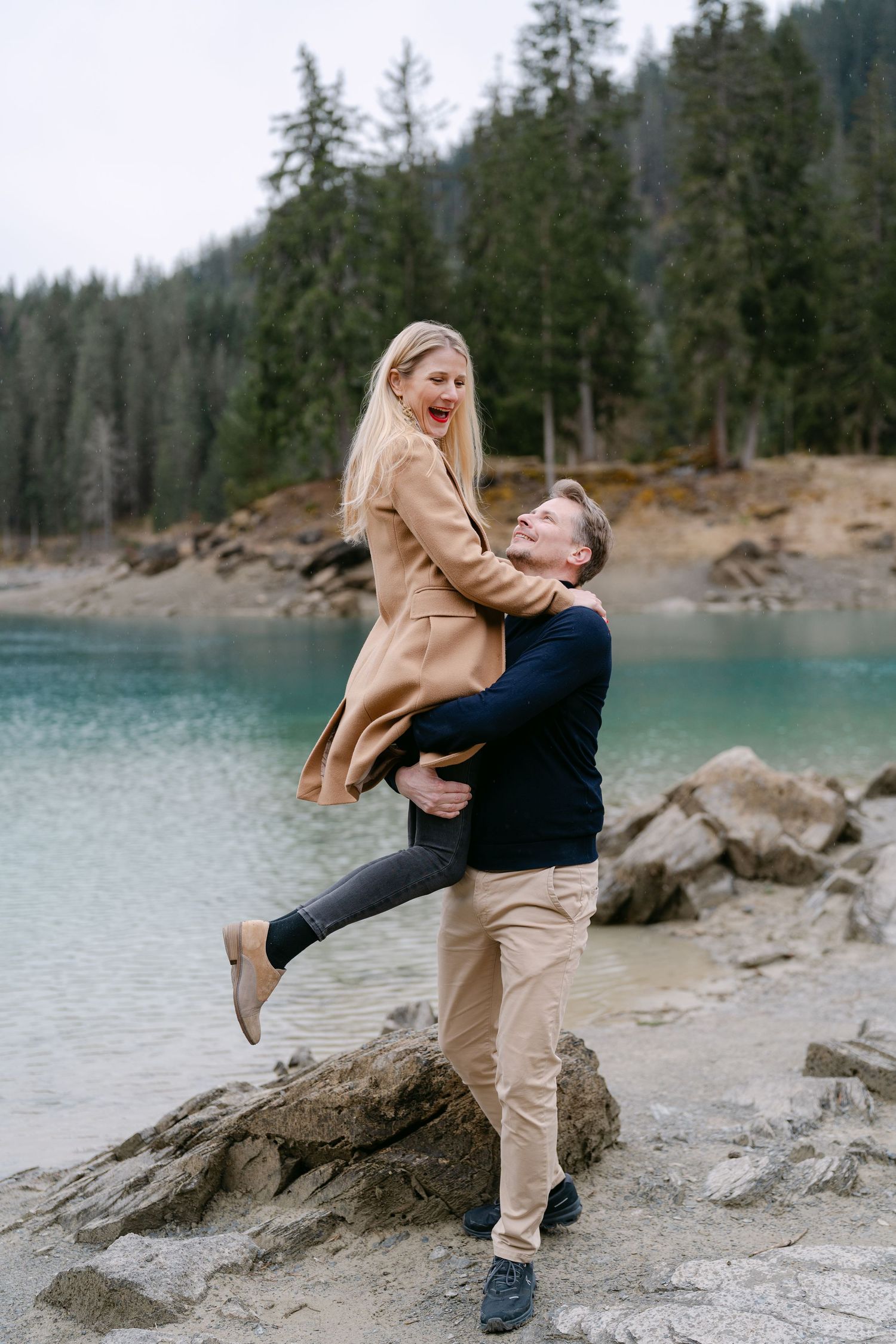 A couple plays and laughs together on rocky shore of turquoise alpine lake.