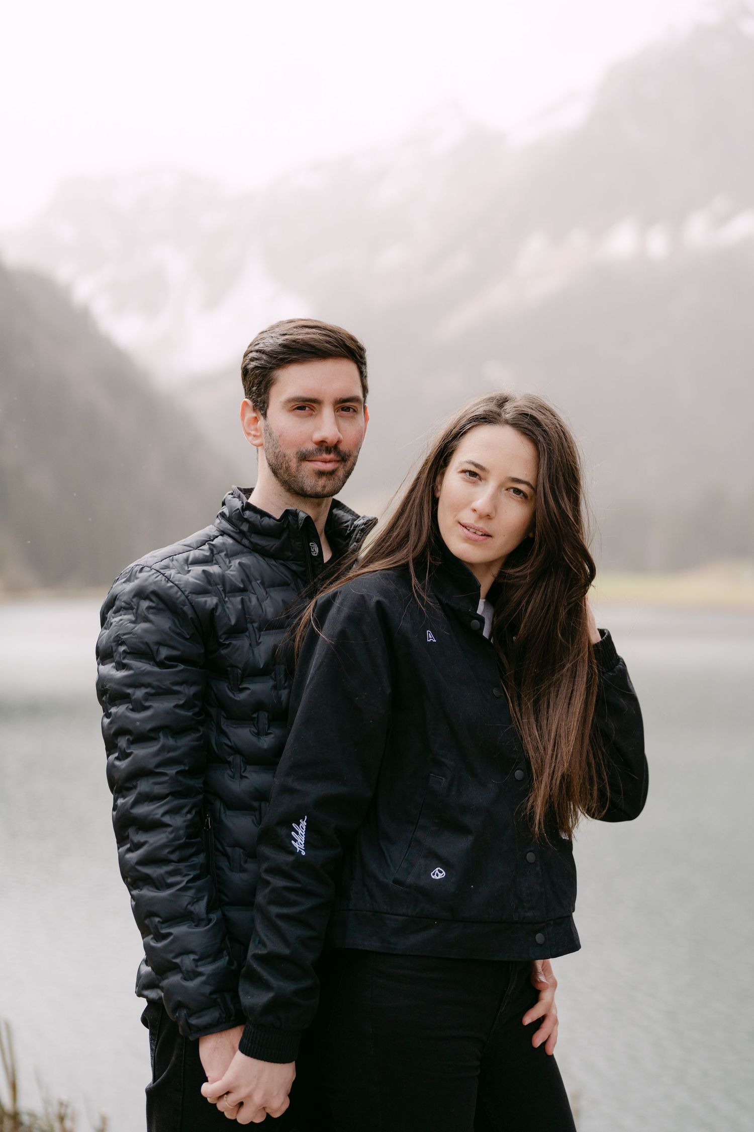 Couple in matching black winter jackets posing by a misty mountain lake.
