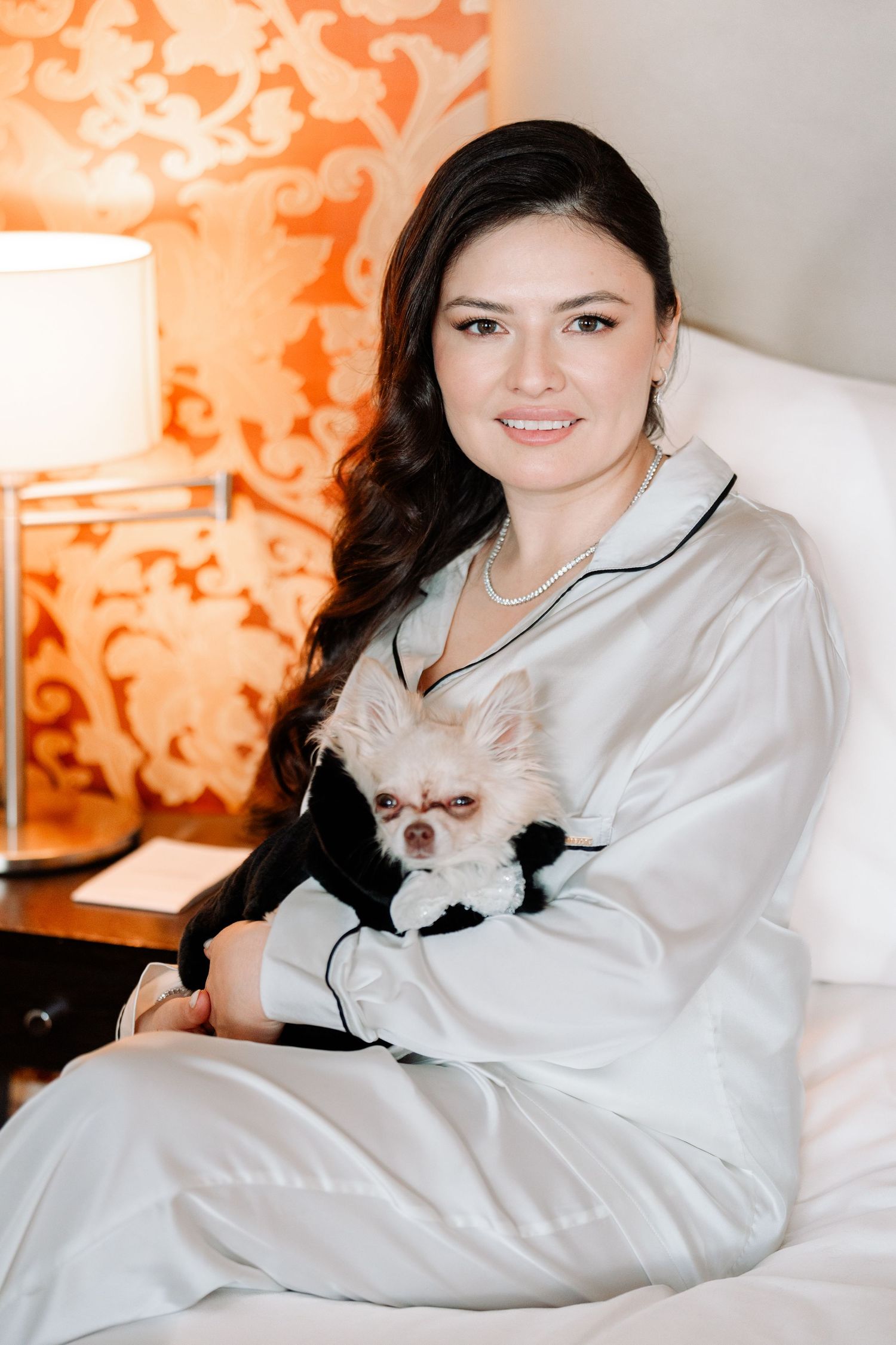 Woman in silk pajamas sits with small Chihuahua dog against orange patterned wallpaper.